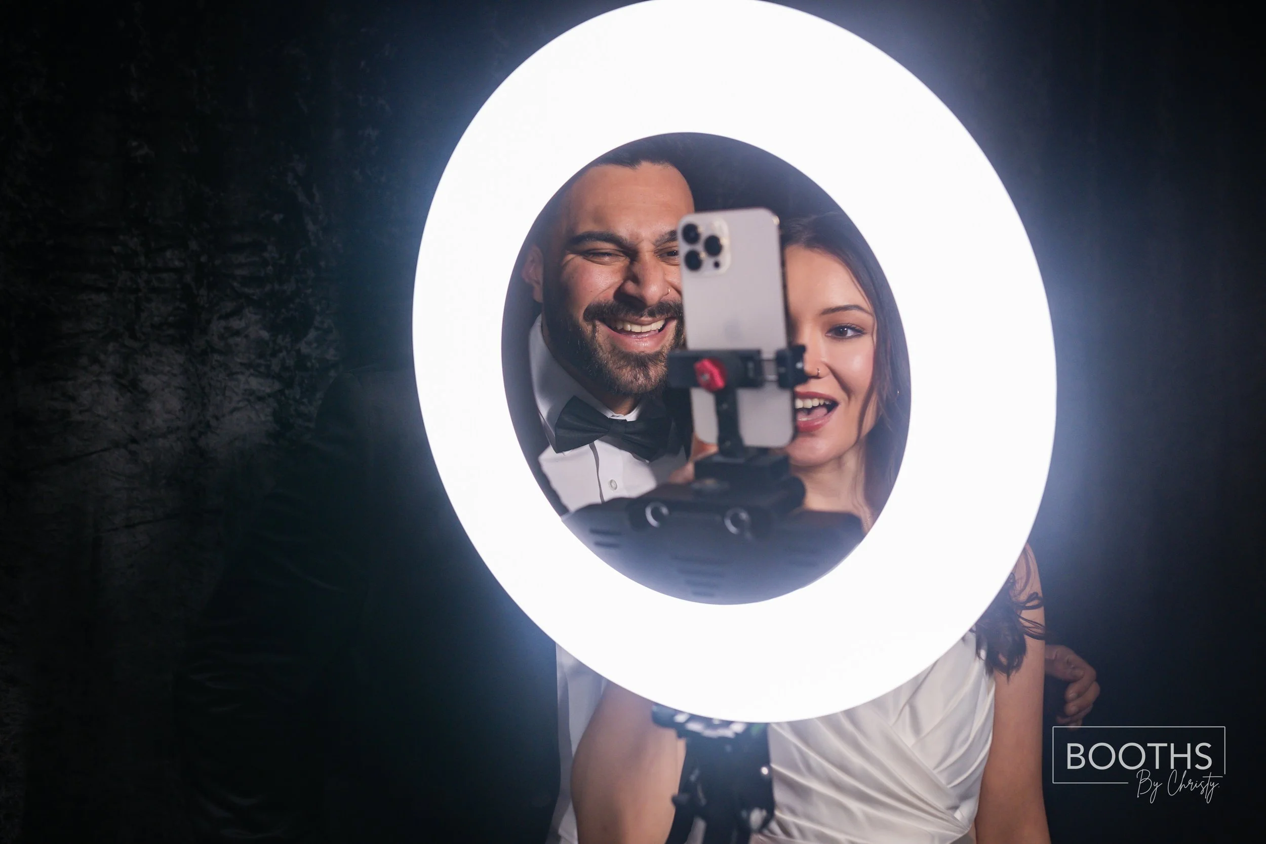 A smiling couple takes a selfie in front of a ring light, dressed formally for an event. The man has a beard and is wearing a tuxedo with a bow tie; the woman has long dark hair and is dressed in a white gown. They are inside a dark room with a brick