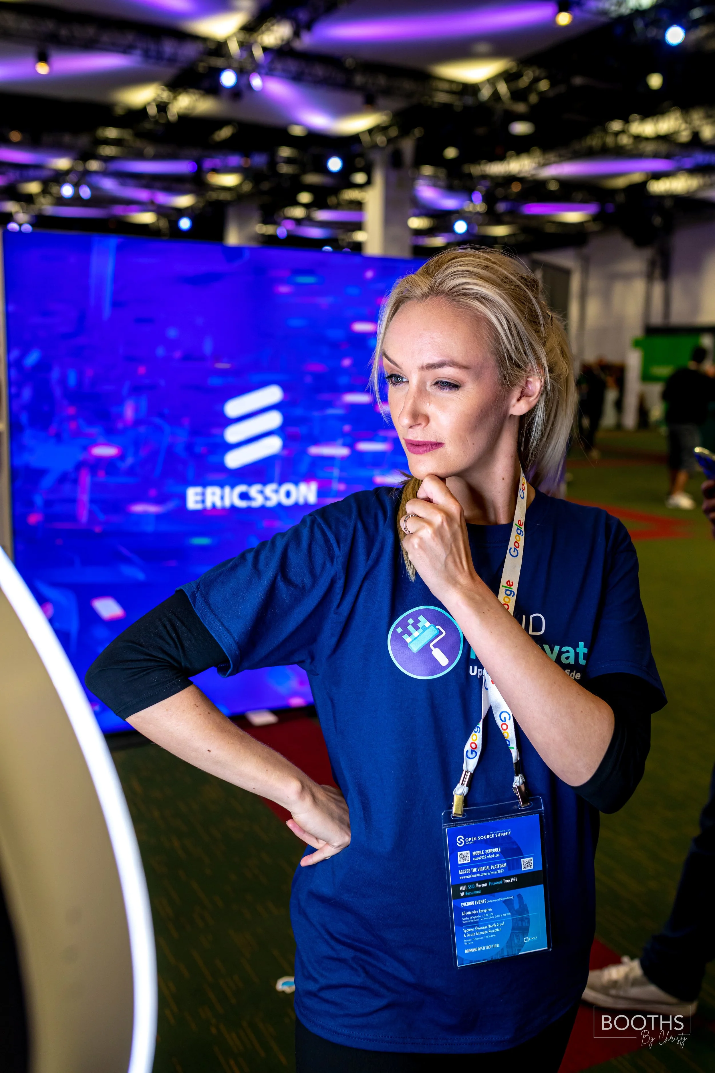 A woman with blonde hair looking thoughtfully at a device at a technology event, wearing a blue t-shirt with a coding icon and a Google lanyard, in front of a large display screen showing the Ericsson logo.