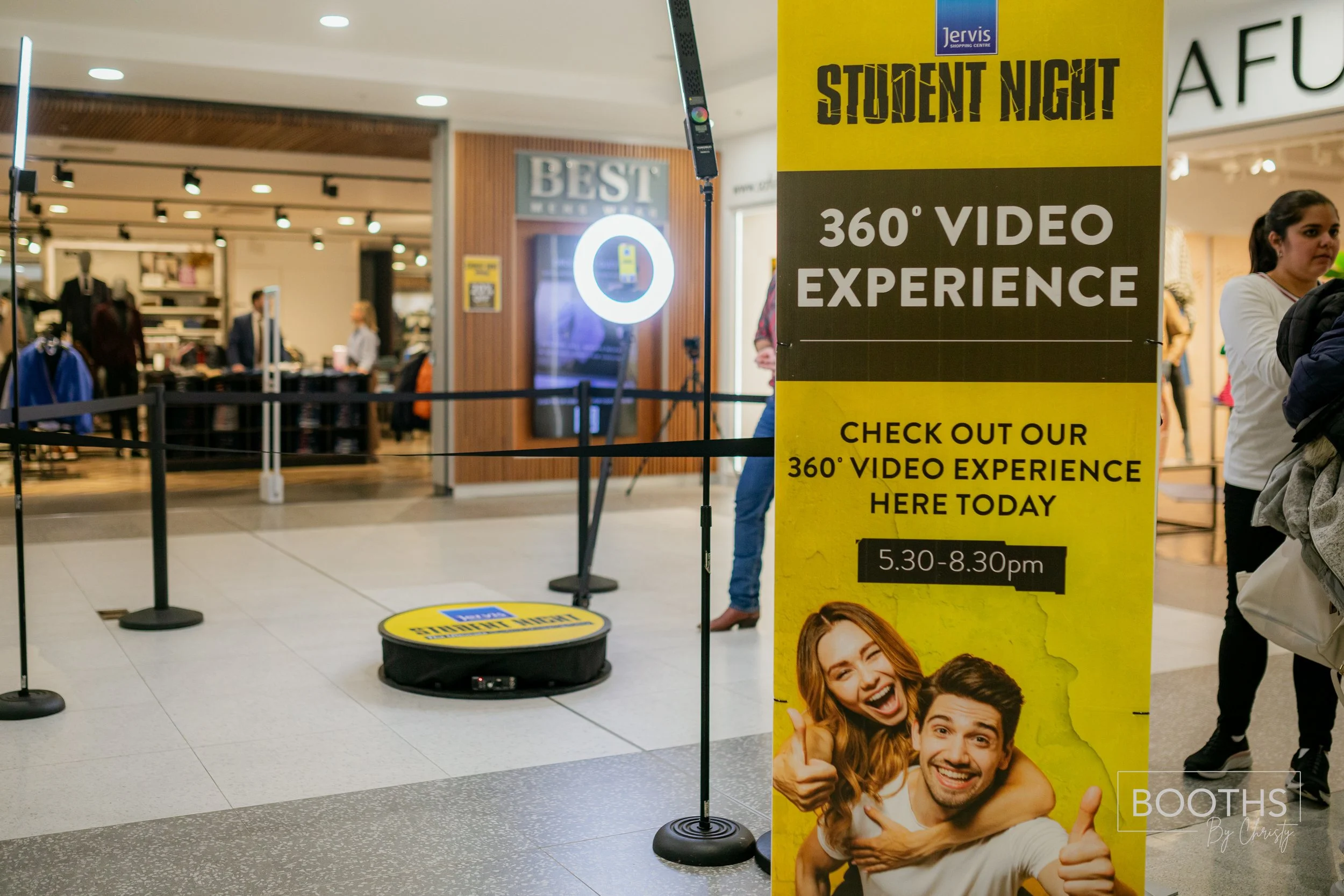 Yellow promotional banner in a shopping mall advertising a student night 360-degree video experience from 5:30 to 8:30 pm. The banner features a young woman and man smiling and giving thumbs up. Behind the banner is a robotic device with a circular y
