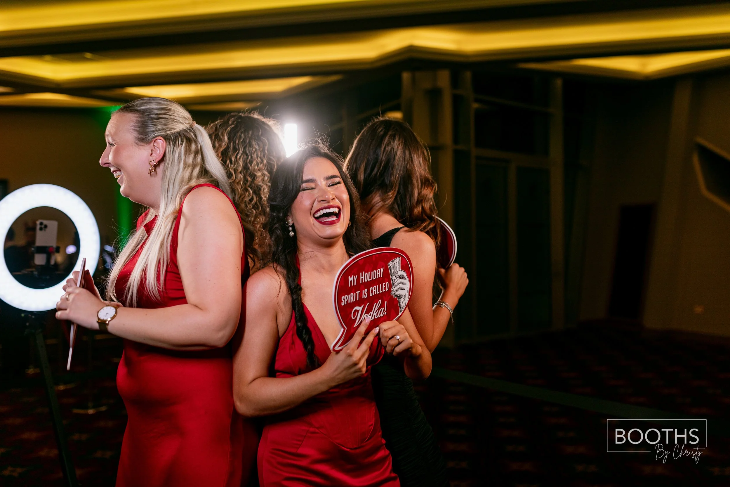 Group of women in red and black dresses celebrating at an event, one woman holding a sign that says 'My holiday spirit is called Vodka!' while laughing.