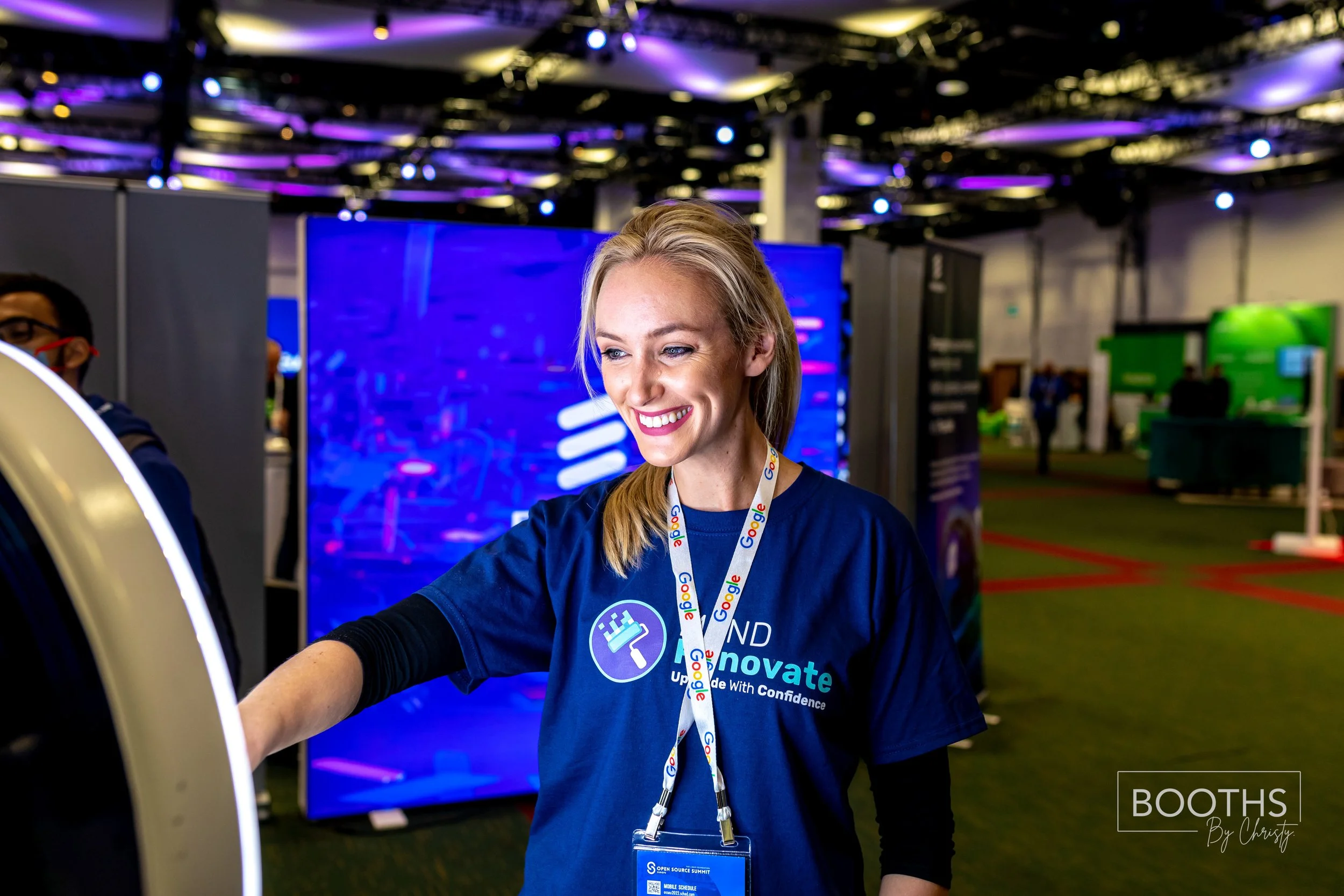Smiling woman wearing a blue t-shirt with the words 'Learn and Innovate' and a badge, taking a photo or selfie at a tech event or conference, with a large digital screen and other attendees in the background.