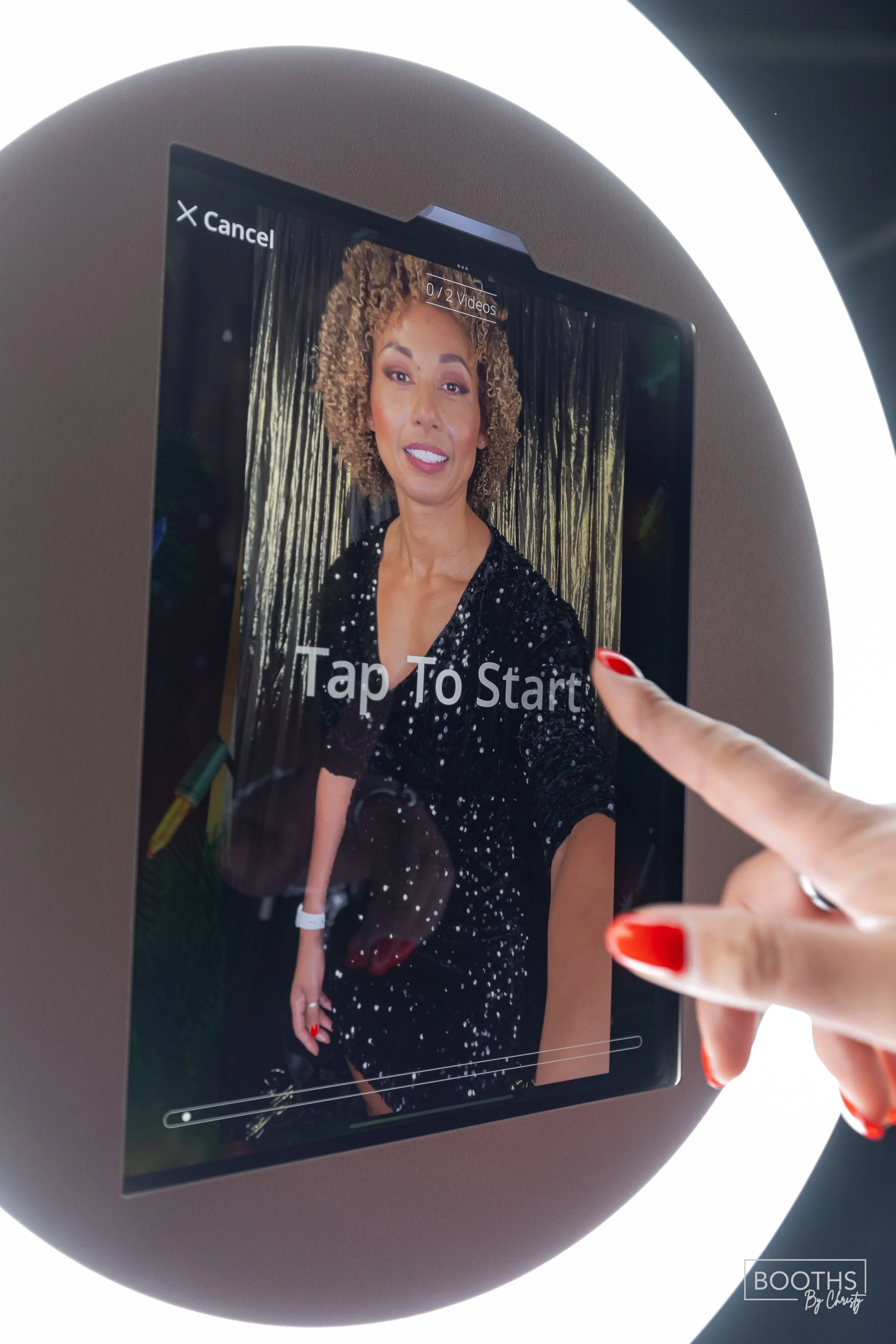 A person with red nail polish is touching a screen that displays a woman with curly hair wearing a black dress with white specks, smiling in front of a dark curtain background.