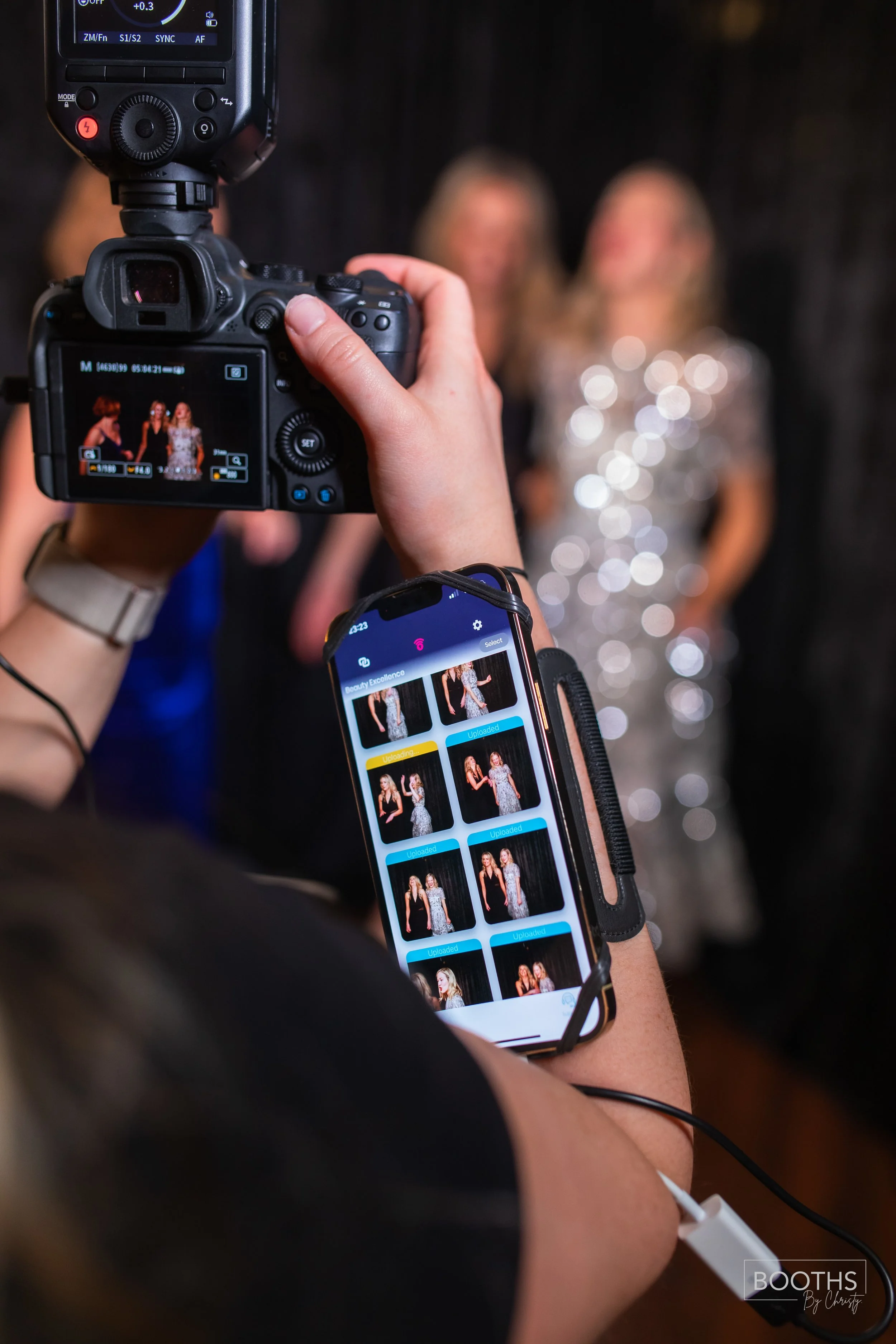 Photographer taking pictures of two women dressed in elegant outfits, with a smartphone showing the shots, against a dark background.