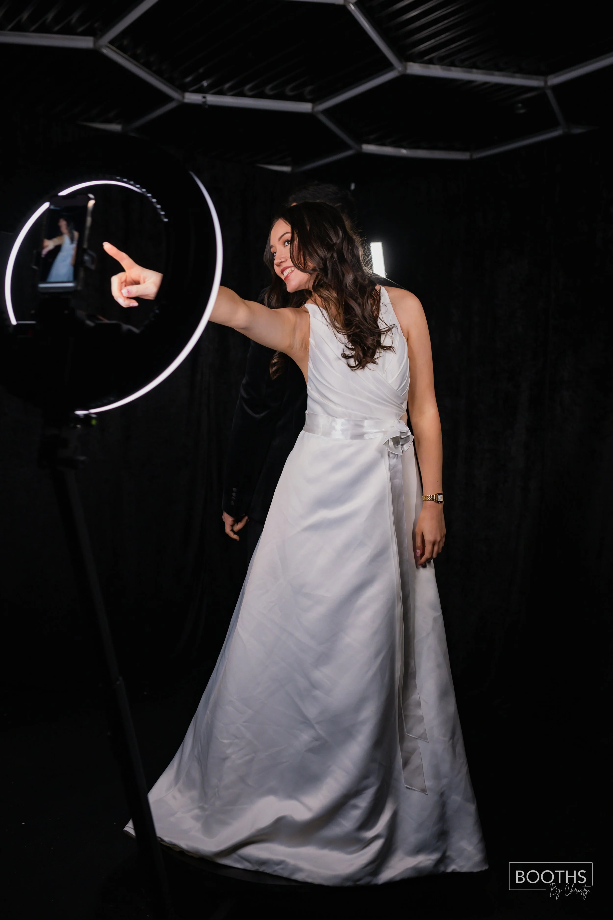 A woman wearing a white wedding dress is taking a selfie with a ring light. She is smiling and pointing at her phone's screen. The background is black.
