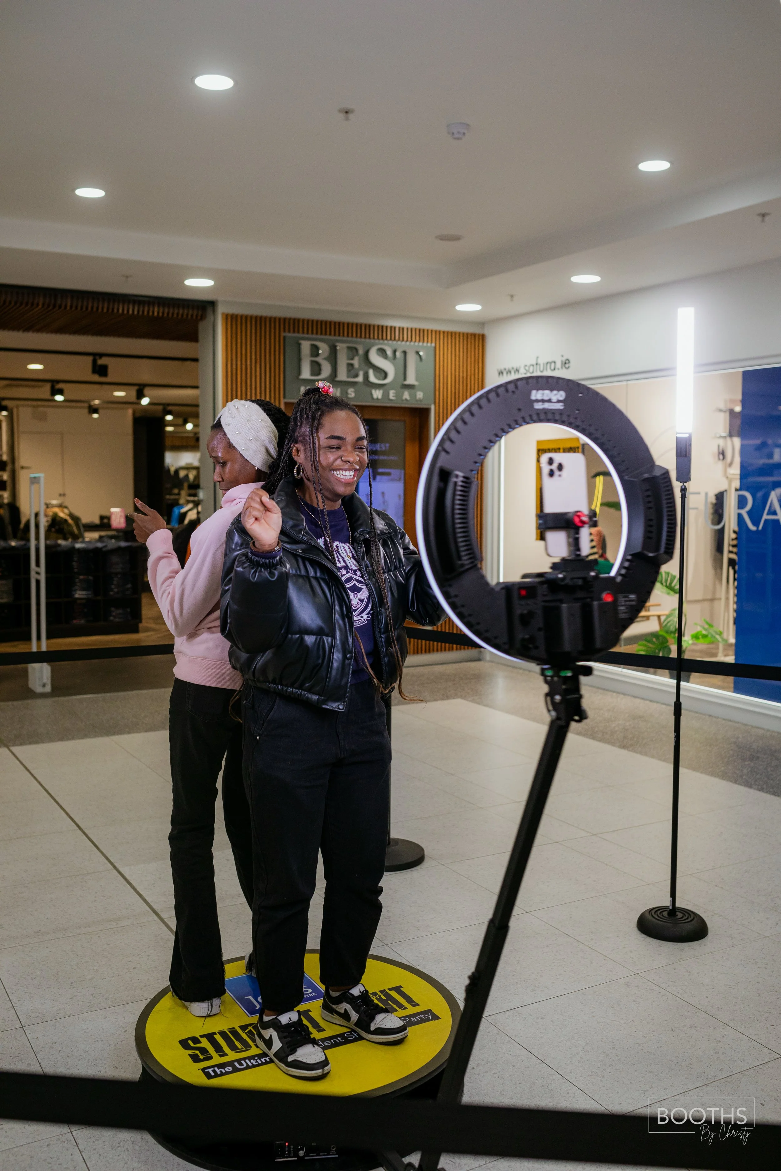 Two women standing in front of a camera setup inside a shopping mall. The woman in front is wearing a black puffer jacket and smiling as she dances, while the woman behind her is looking at her phone. The shopping mall background includes stores and