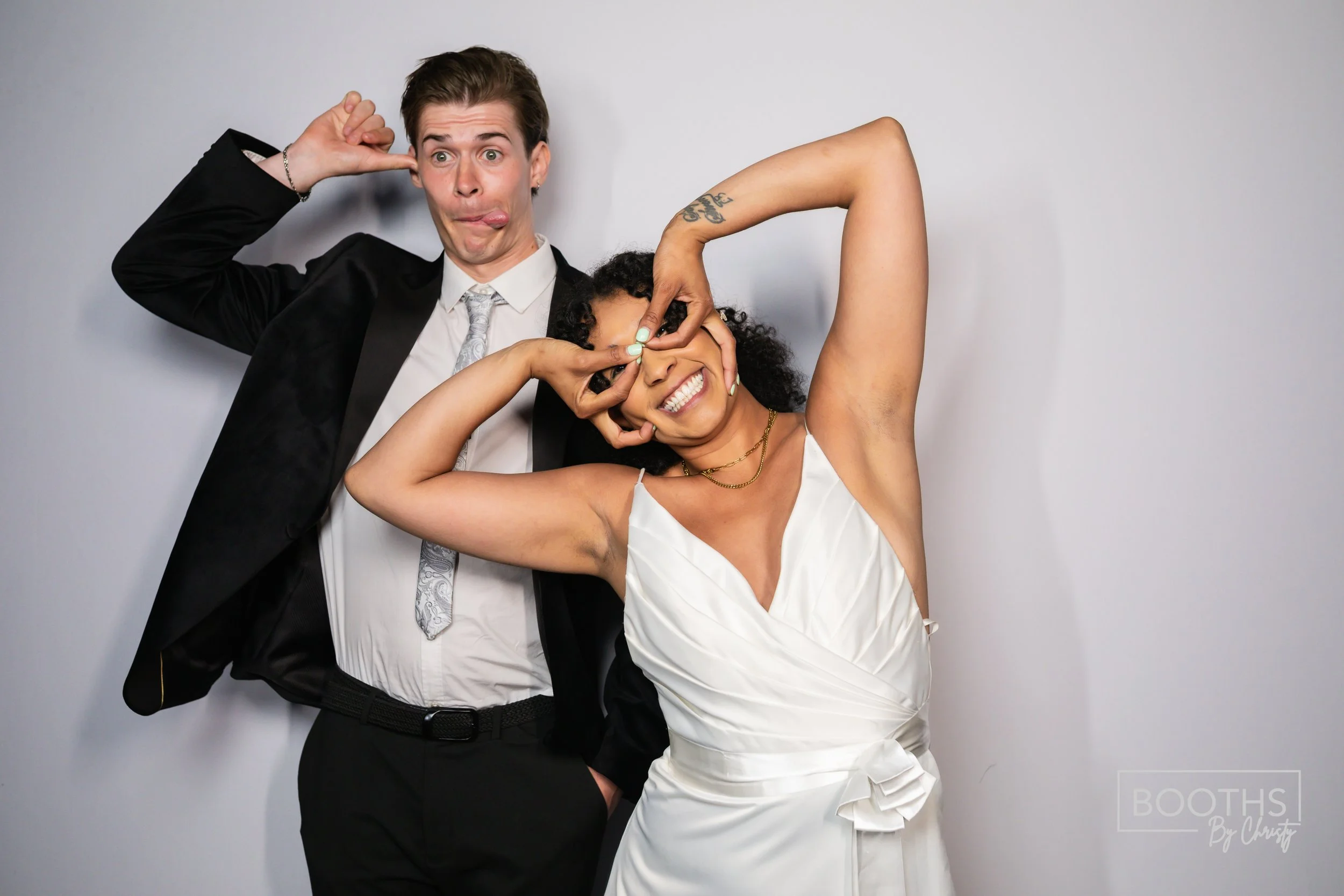 Two friends, a man and a woman, playful and goofy expressions posing together in front of a plain gray backdrop. The man is wearing a black tuxedo with a white shirt and a patterned tie, sticking his tongue out and making a funny face. The woman is w