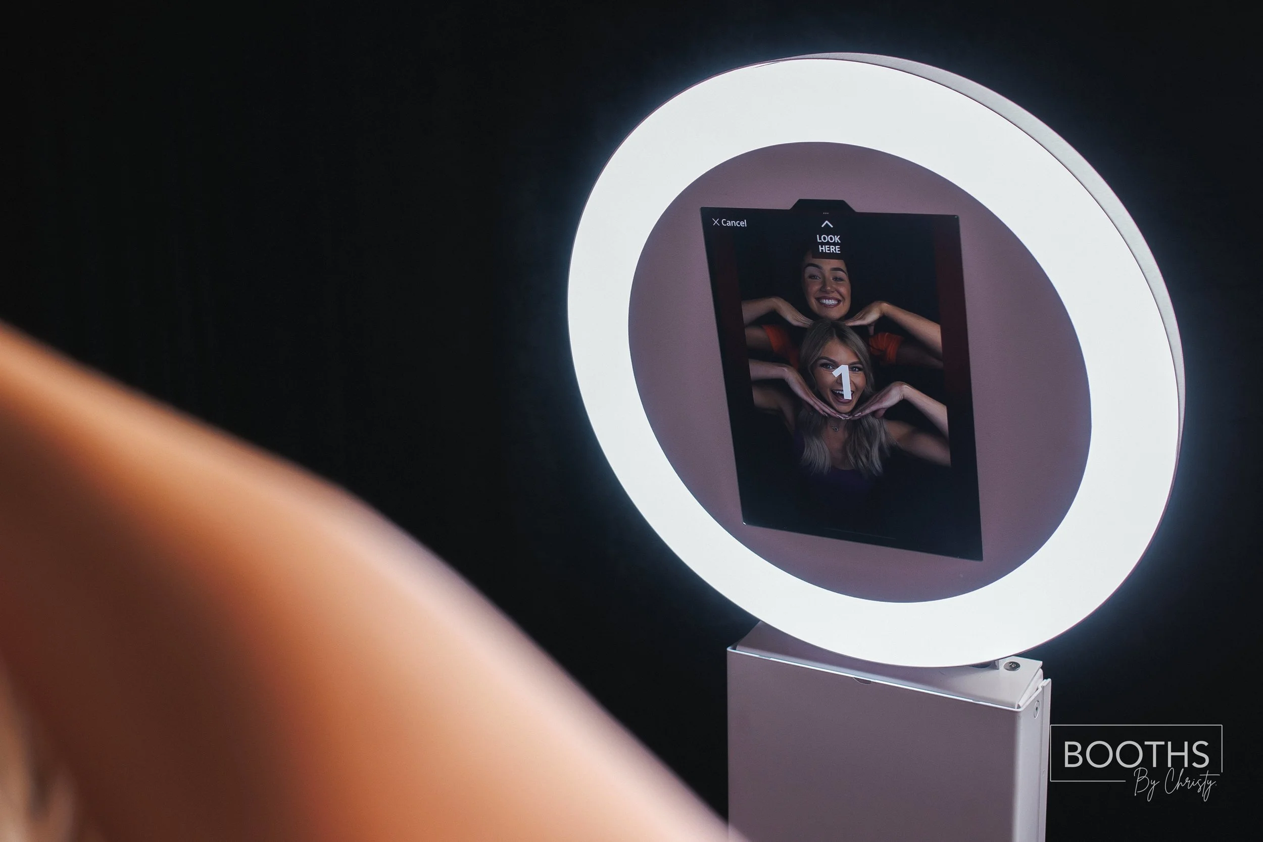 Photo of a photo booth with a ring light around it, showing a digital photo of three women making fun poses. One woman is smiling broadly, the second has her hands framing her face, and the third is partially visible, all with playful expressions.