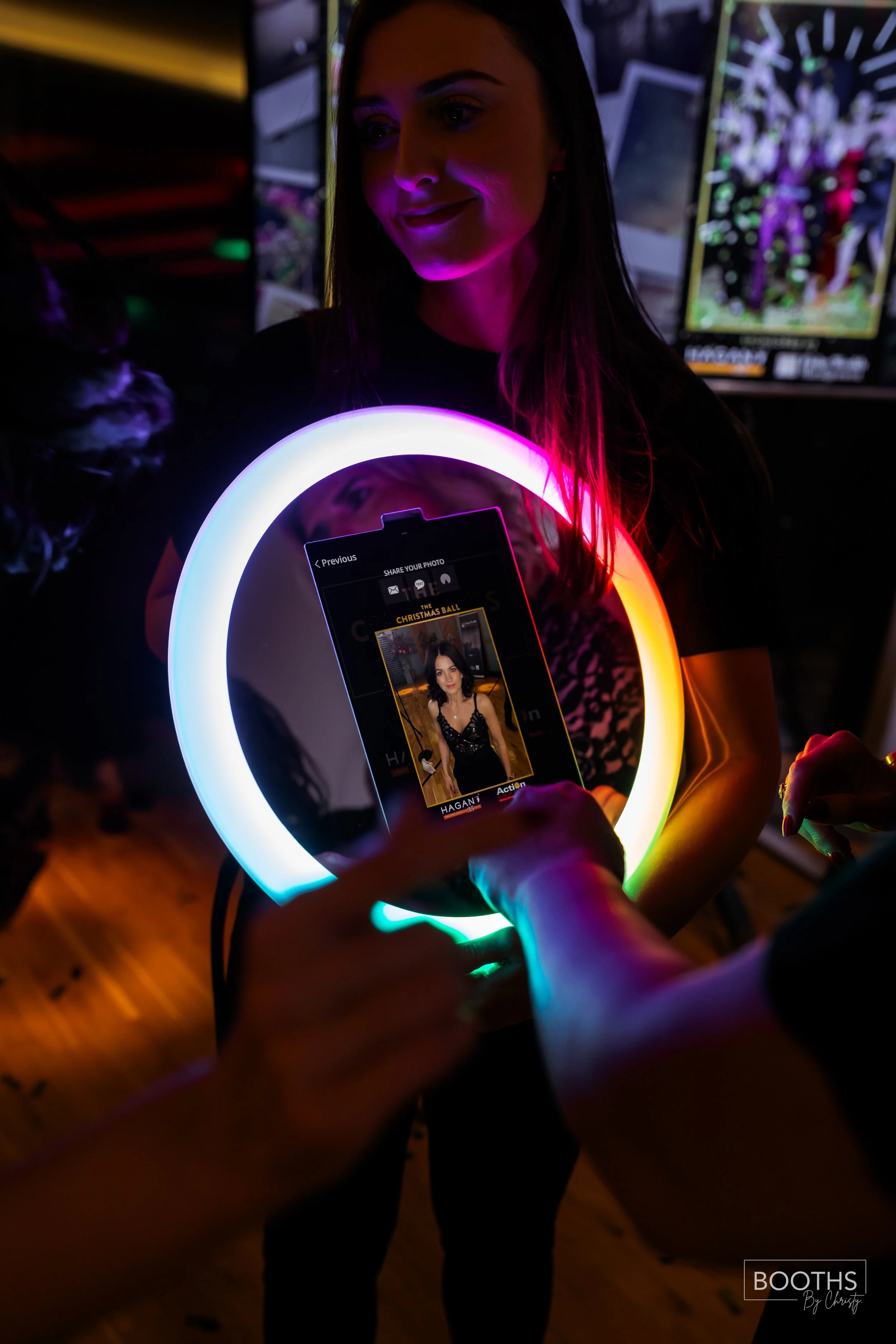 A woman is posing for a photo at a dimly lit event, illuminated by a colorful LED ring light. She is looking at a phone displaying her picture, and someone is taking the photo. There are screens with digital content in the background, and the scene h