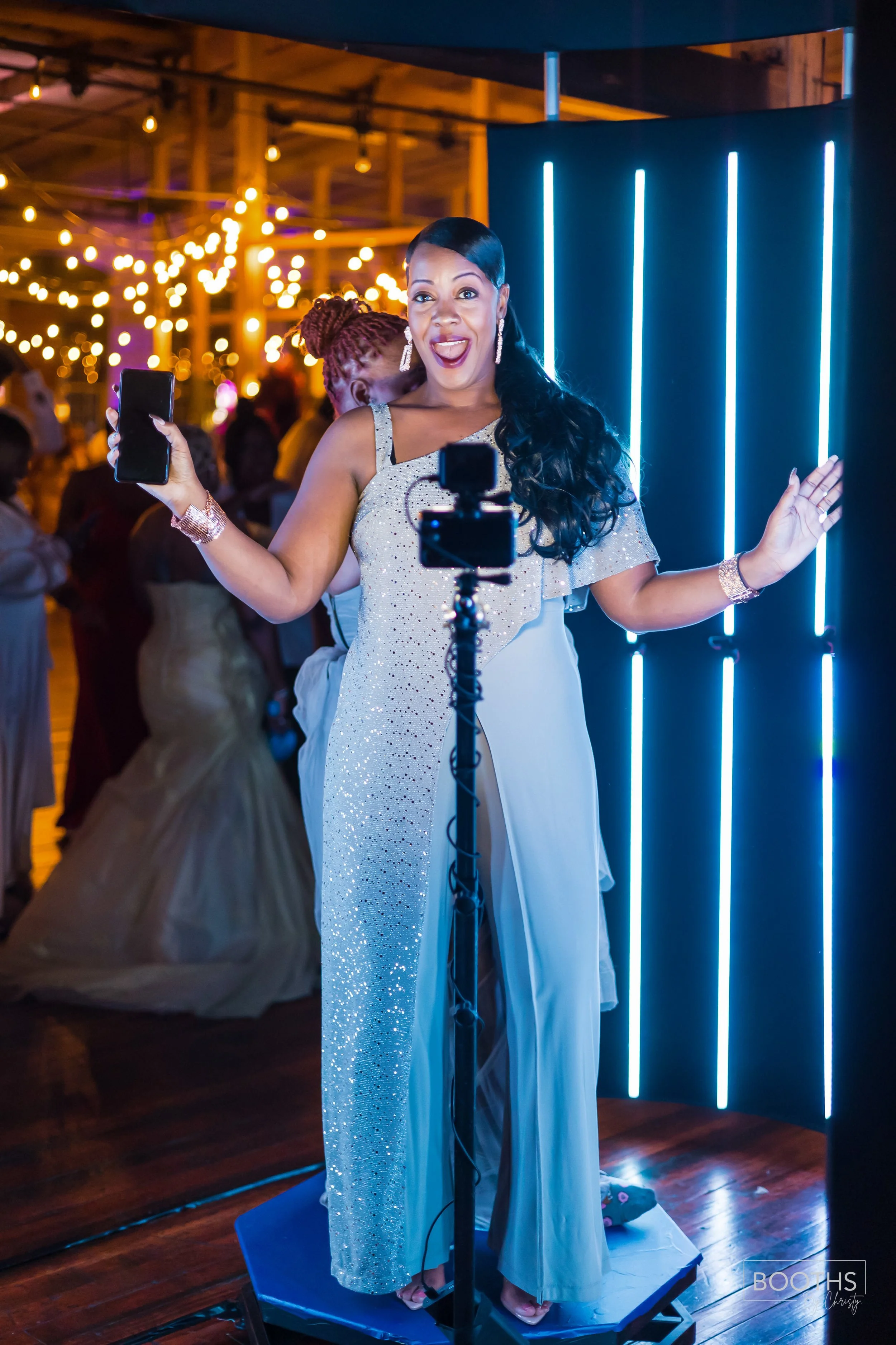 A woman in a sparkly dress taking a selfie at a celebration or party, with a DJ or photographer setup in front of her and guests dancing in the background, illuminated by warm string lights.