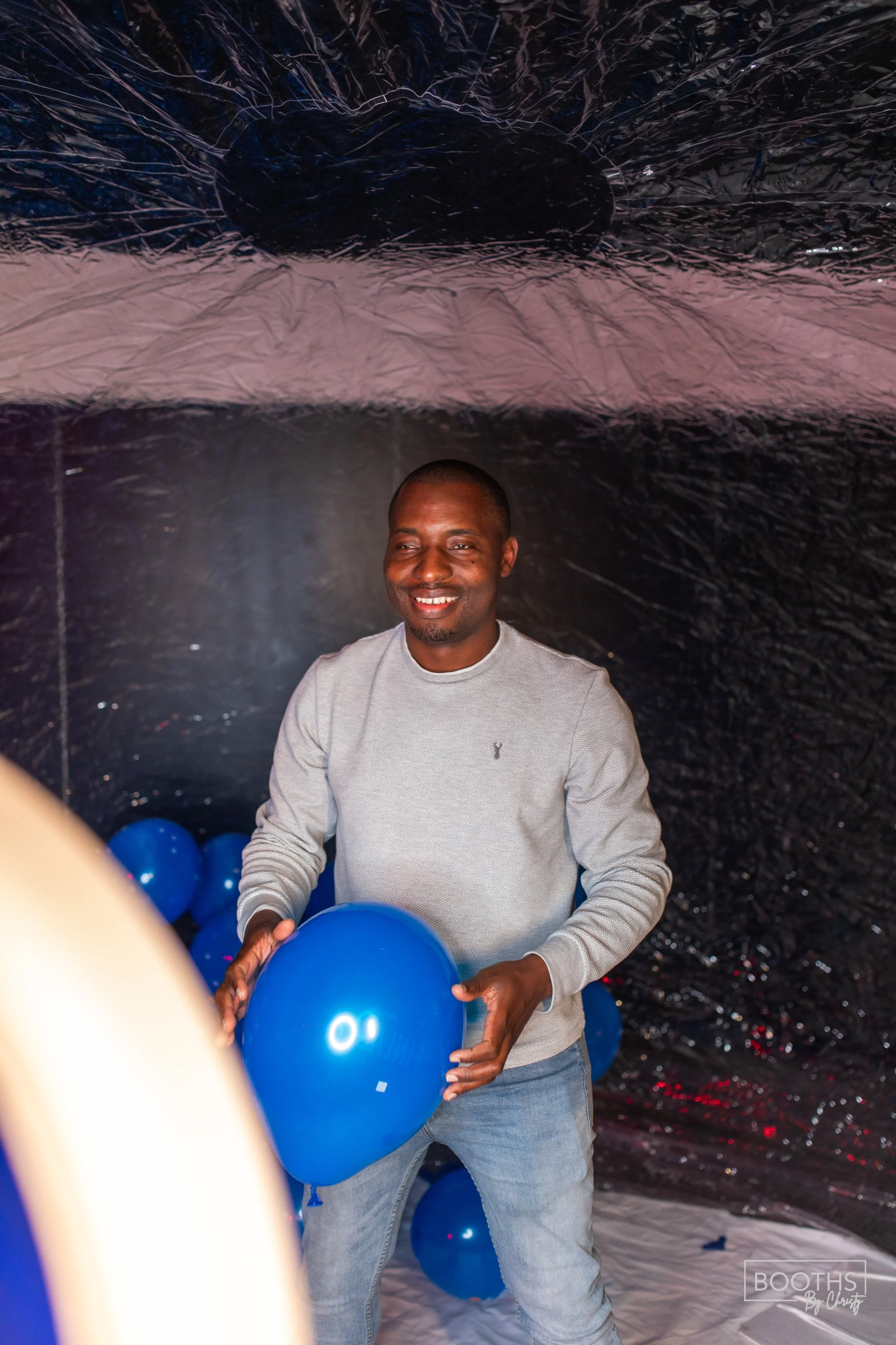 A man smiling and holding blue balloons inside a space decorated with more blue balloons and black plastic background.