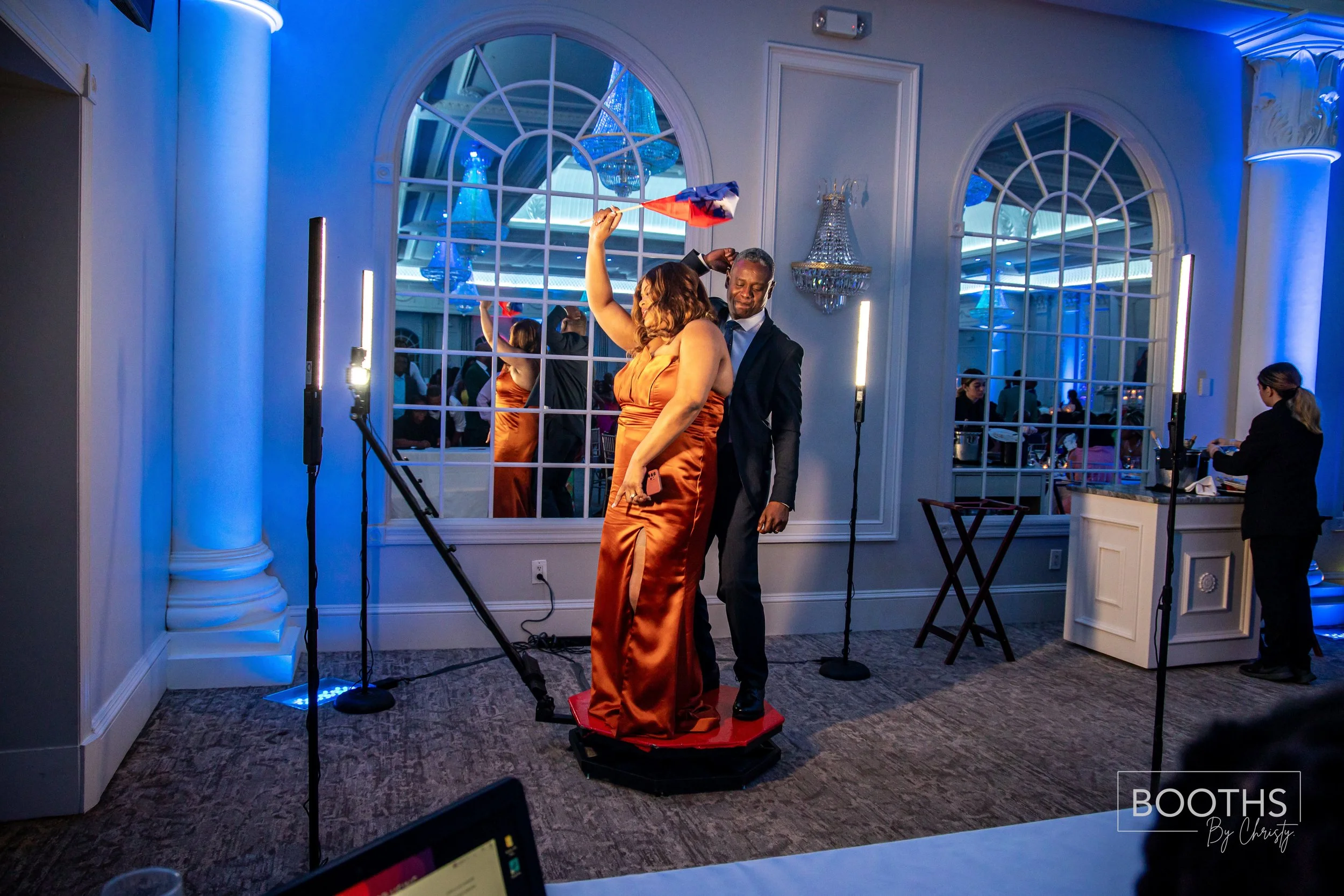 A woman in a shiny orange gown and a man in a black suit and tie dancing together on a small red platform at a celebration event, with a woman in black serving at a table in the background.