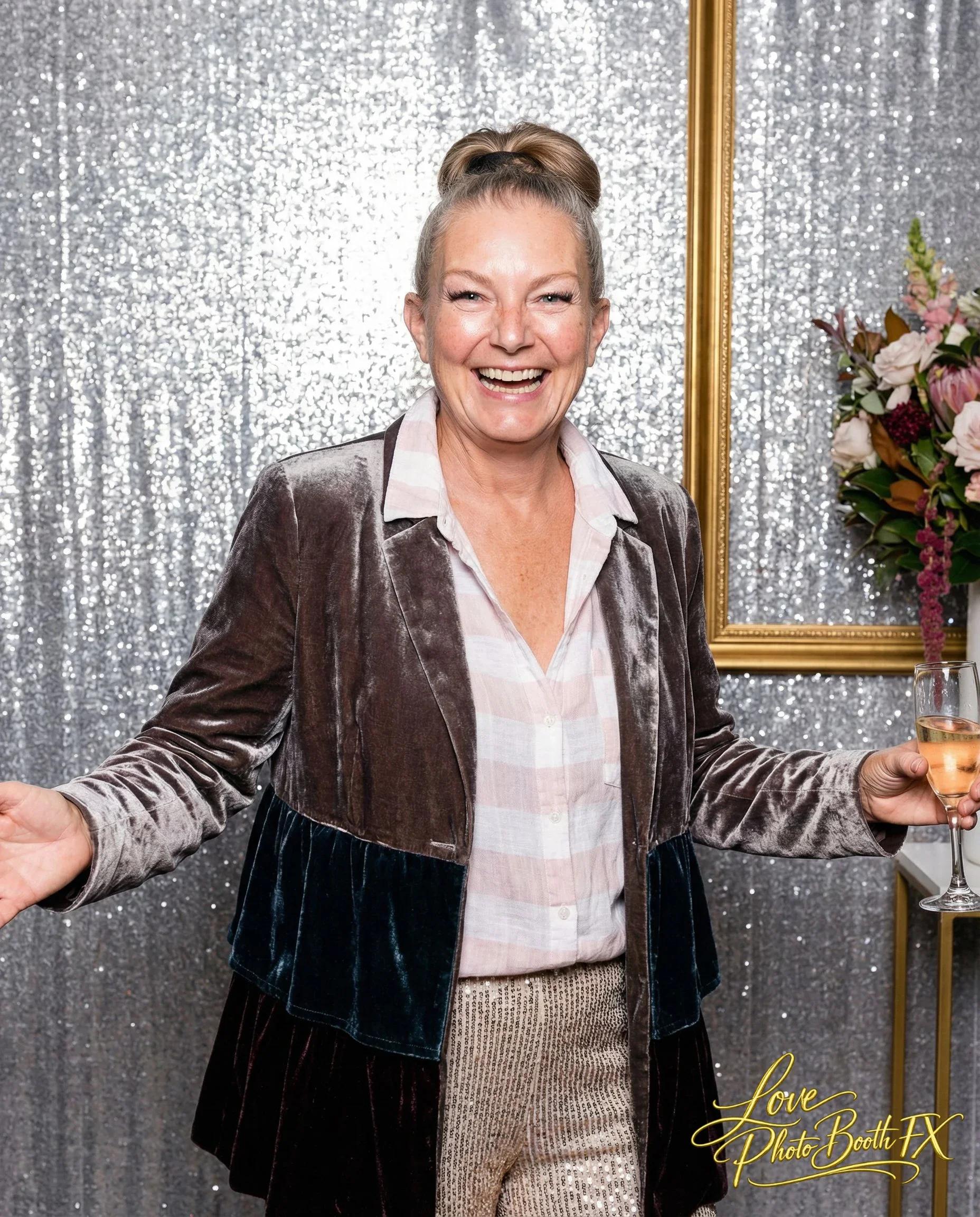 A smiling woman holding a glass of champagne at a photo booth with a sparkly silver background and a floral arrangement to the side.
