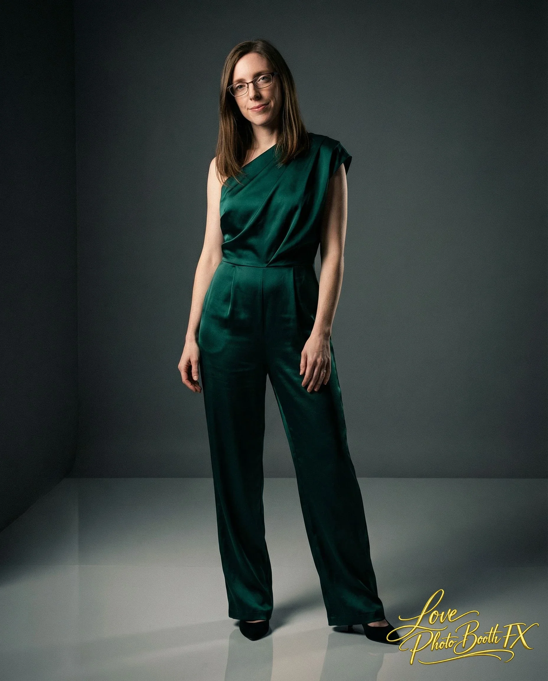 A woman with brown hair, glasses, and a slight smile, wearing a dark green satin sleeveless jumpsuit, standing in a gray photo studio with a dark background.