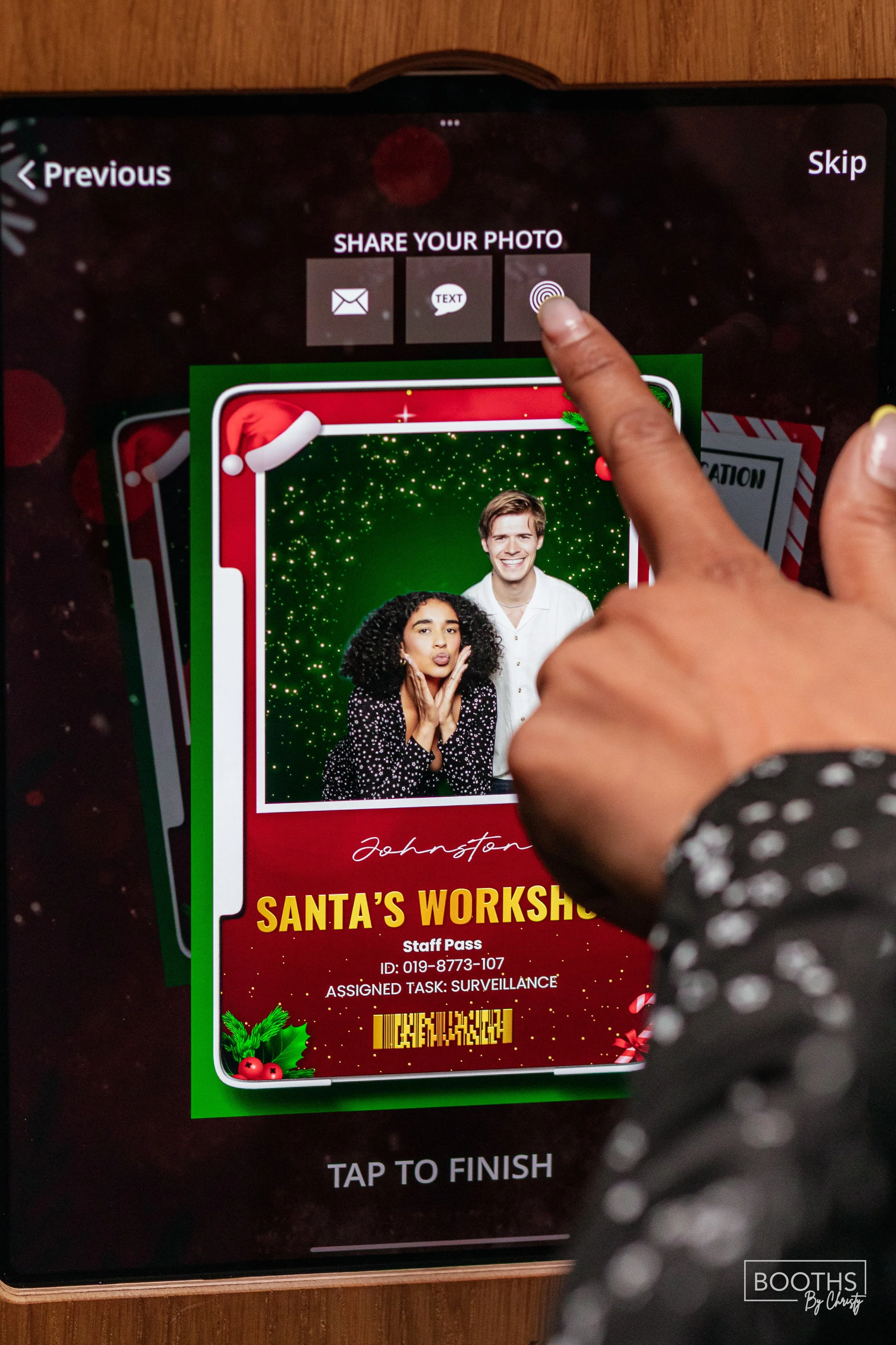 Touchscreen at a Christmas-themed photo booth with a digital photo of a woman and a man in front of a green sparkly background, and a printed holiday pass reading "Santa's Workshop" with a barcode, staff ID, and surveillance task.
