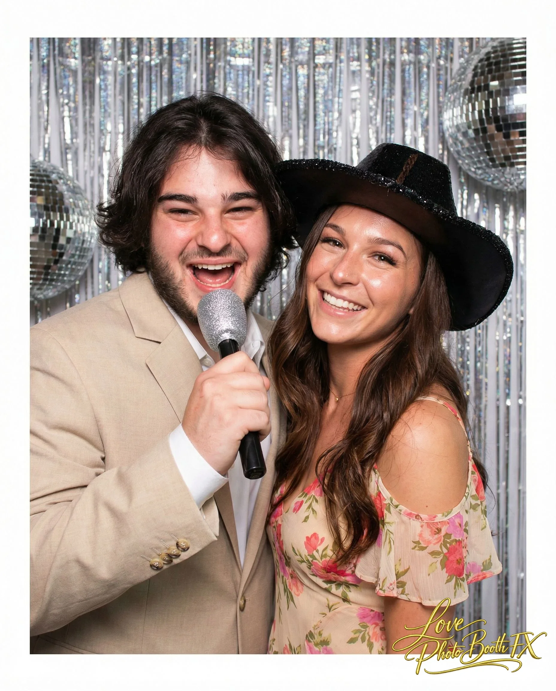 A man and a woman smiling at a photo booth, holding a microphone, with silver tinsel and disco balls in the background. The woman is wearing a floral dress and a wide black hat, the man is in a beige suit.