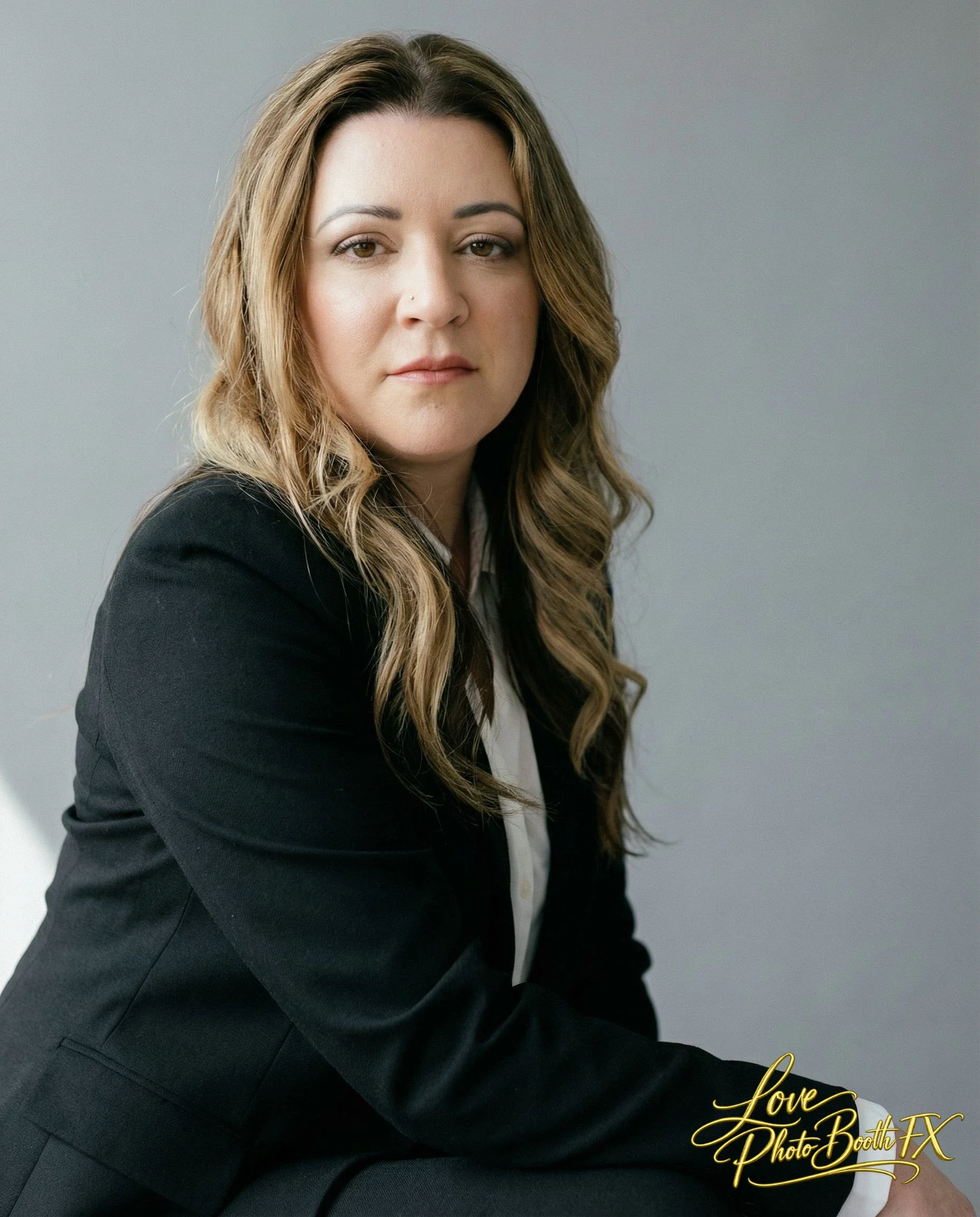 A woman with long wavy hair wearing a black blazer and white shirt, seated against a gray background.