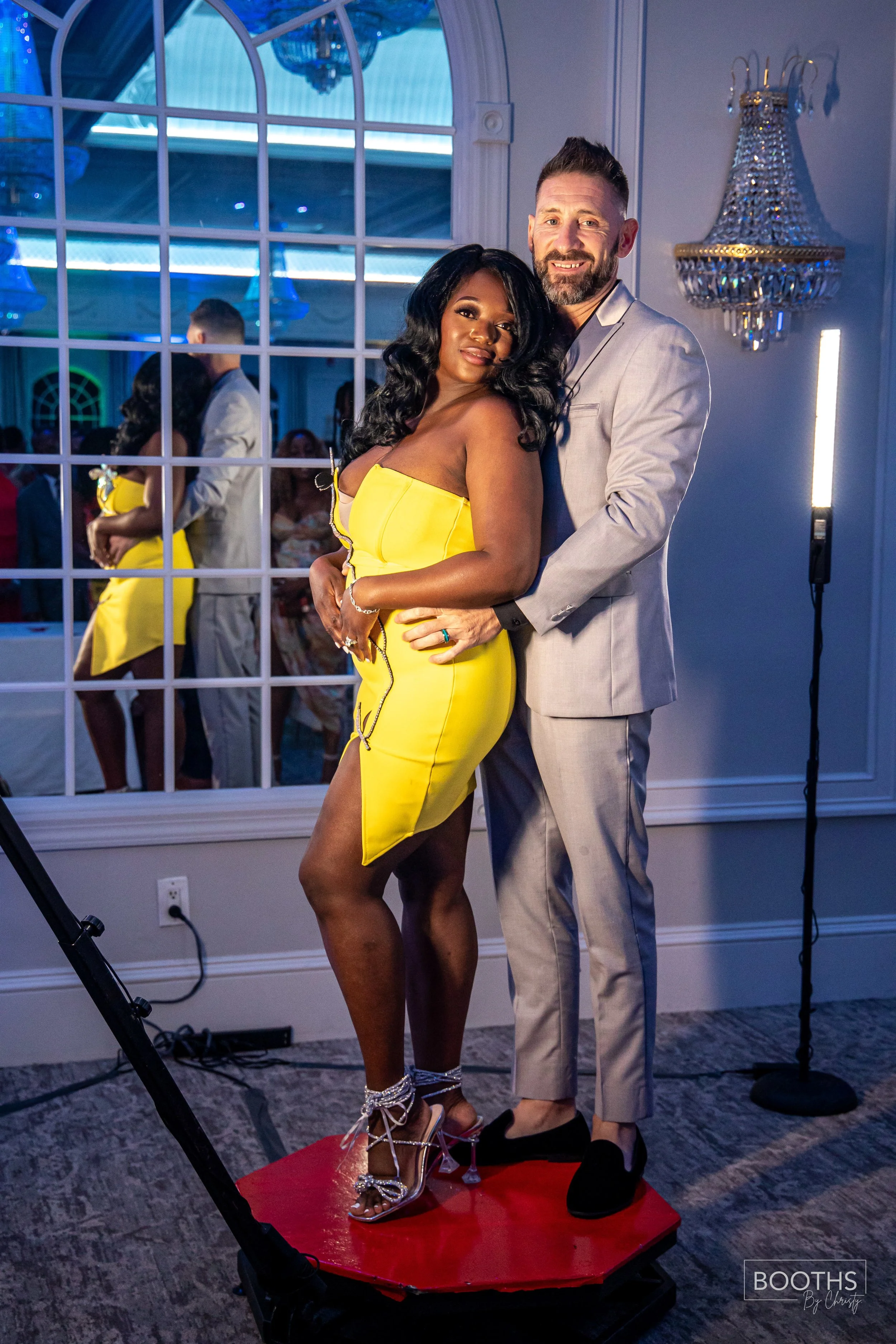 A couple dressed in formal attire, standing close together on a red platform at an indoor event, with the woman in a yellow dress and the man in a light gray suit, smiling at the camera.