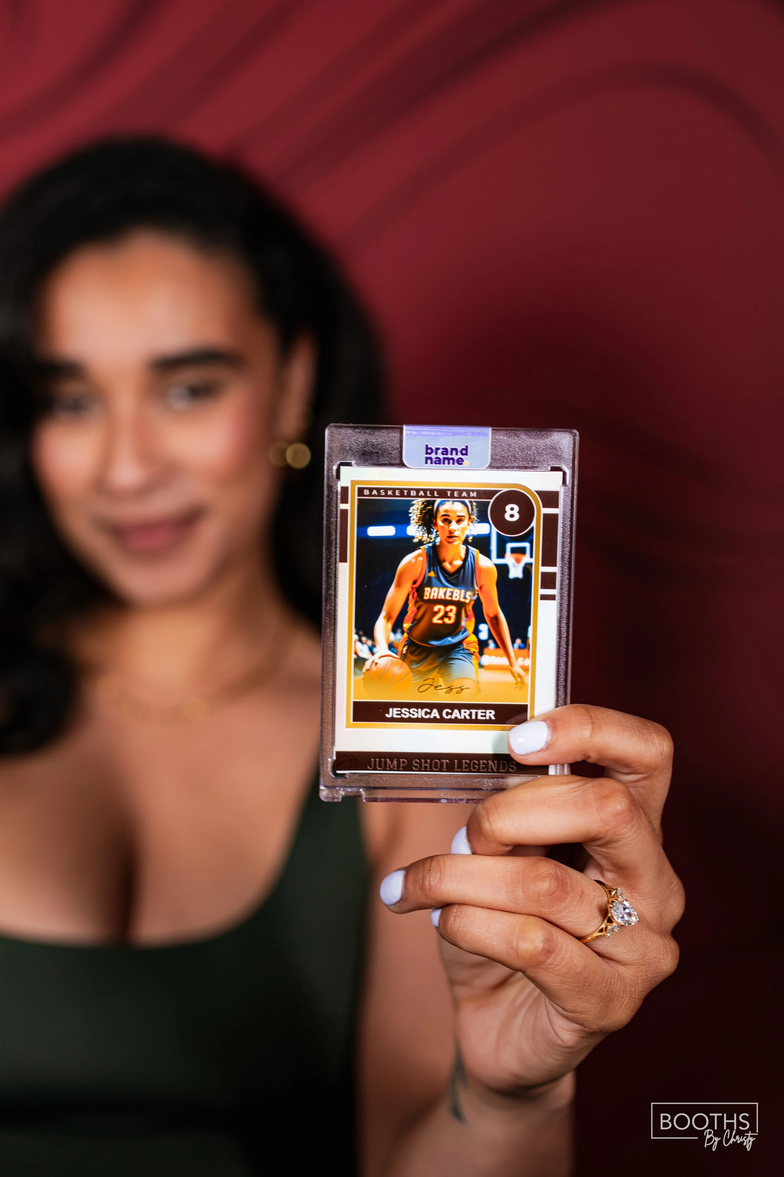A woman with dark hair and a dark green top holding a basketball card of Jessica Carter, wearing a basketball uniform, in front of a blurred pink background.