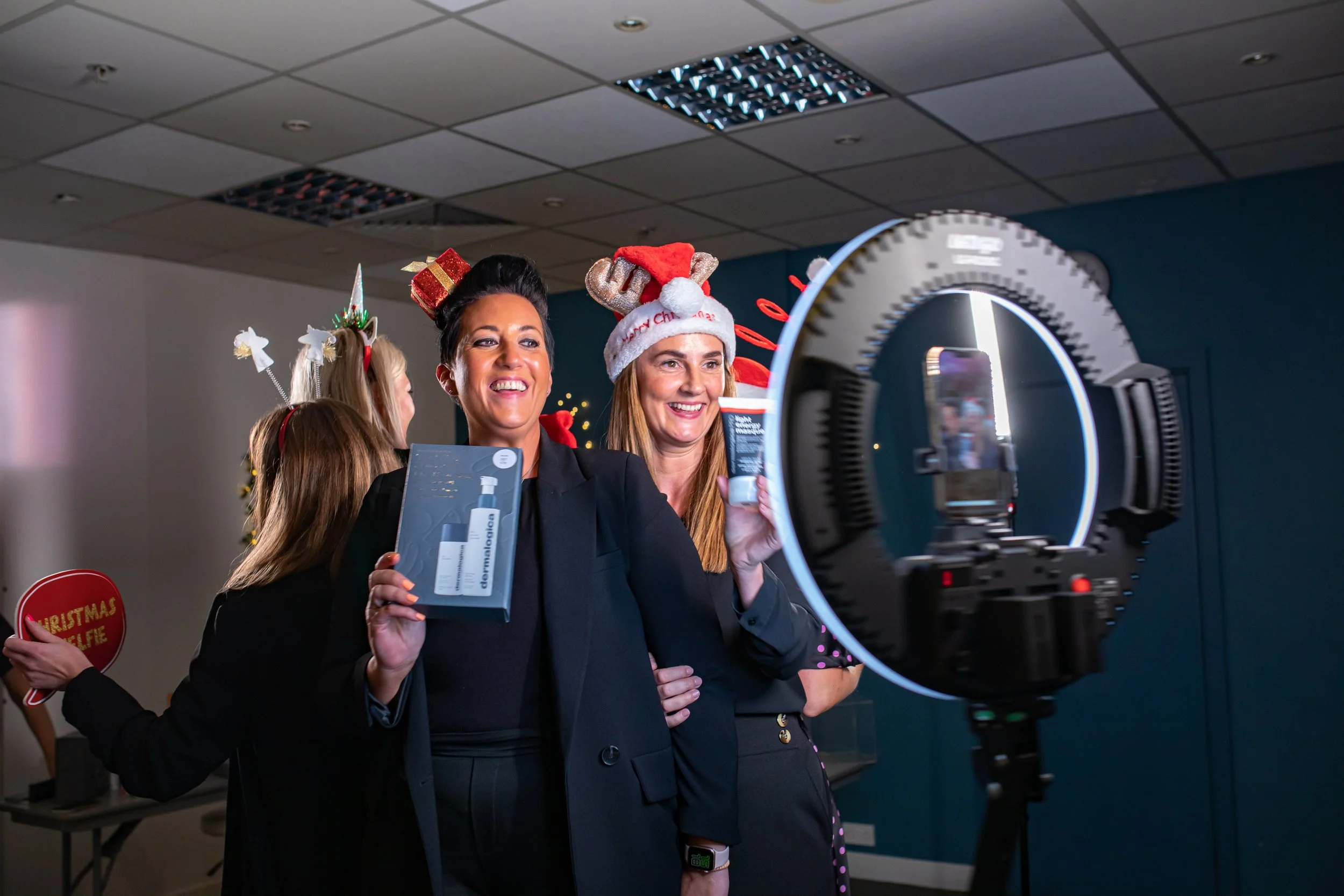Women celebrating Christmas, taking a selfie with a ring light, wearing festive hats and holding beauty product boxes, in an office decorated for Christmas.