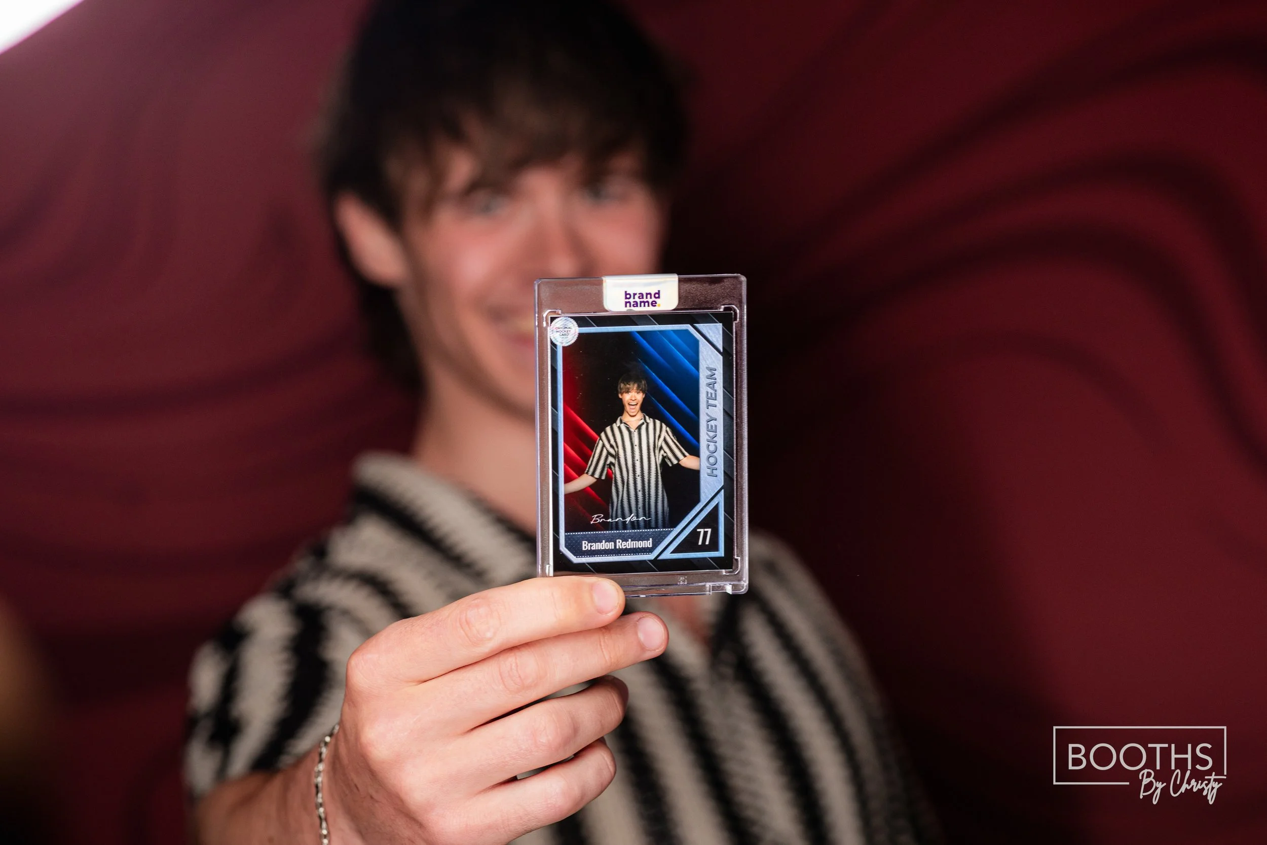 A person holding up a hockey team ID badge with a photo of a young man in a black and white striped referee shirt, tan skin, and short dark hair, against a red background.