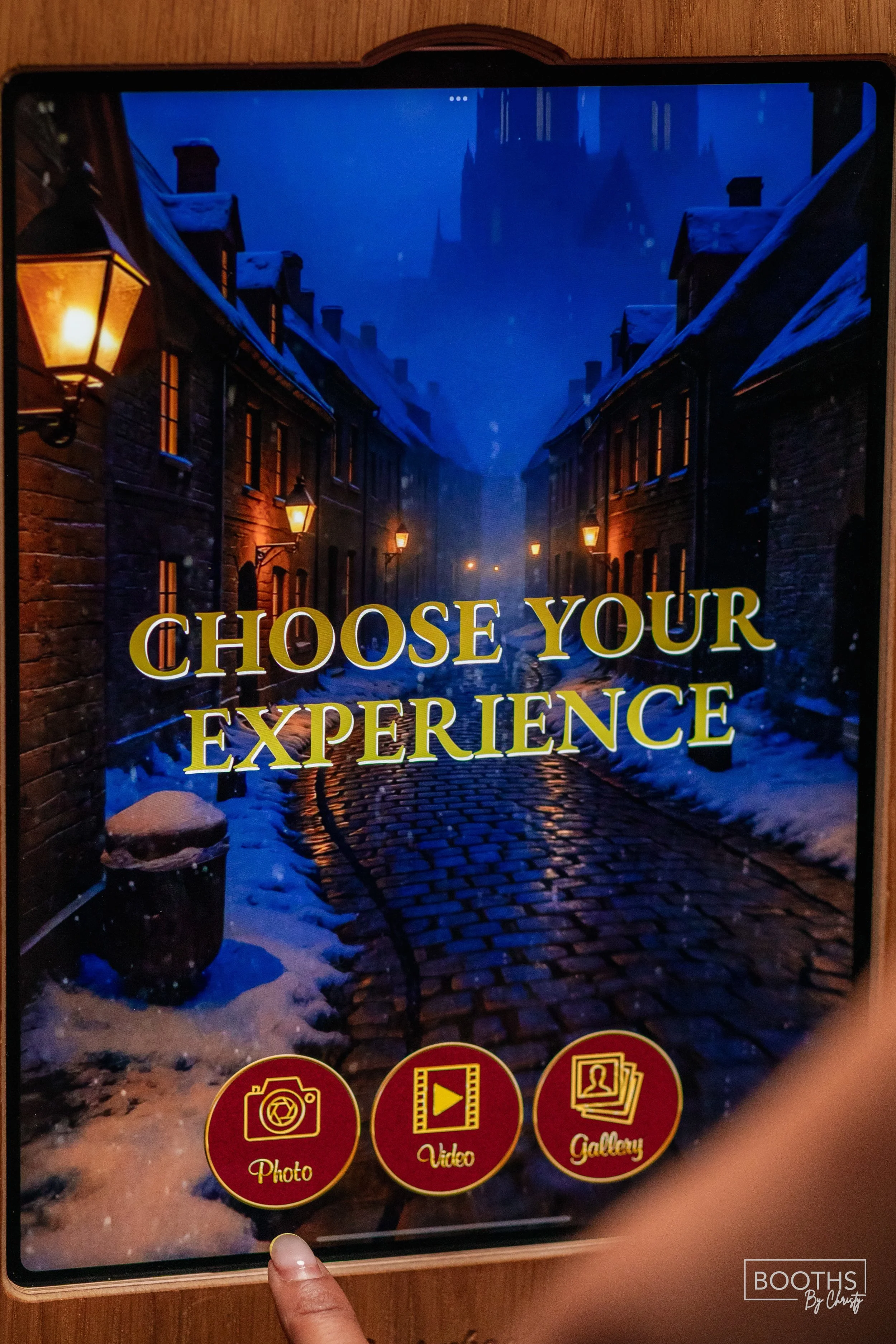 An interactive kiosk screen with a background of a snowy, cobblestone street lit by street lamps at dusk, featuring the phrase "Choose Your Experience" and three red buttons labeled Photo, Video, and Gallery.