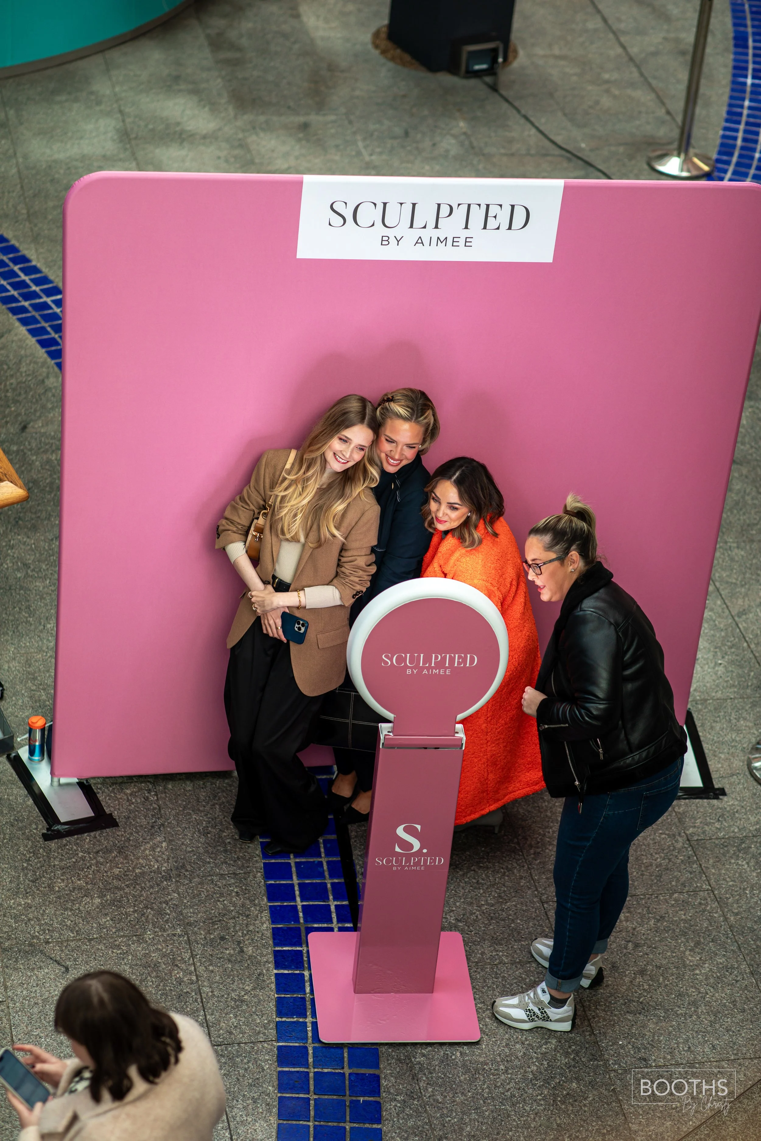 Four women posing for a photo at a pink display booth for 'Sculpted by Aimee'. They are standing close together with two women holding a smartphone, one woman wearing glasses and a black leather jacket, and one woman wrapped in an orange blanket. The booth has a pink backdrop with the logo 'SCULPTED BY AIME' at the top.