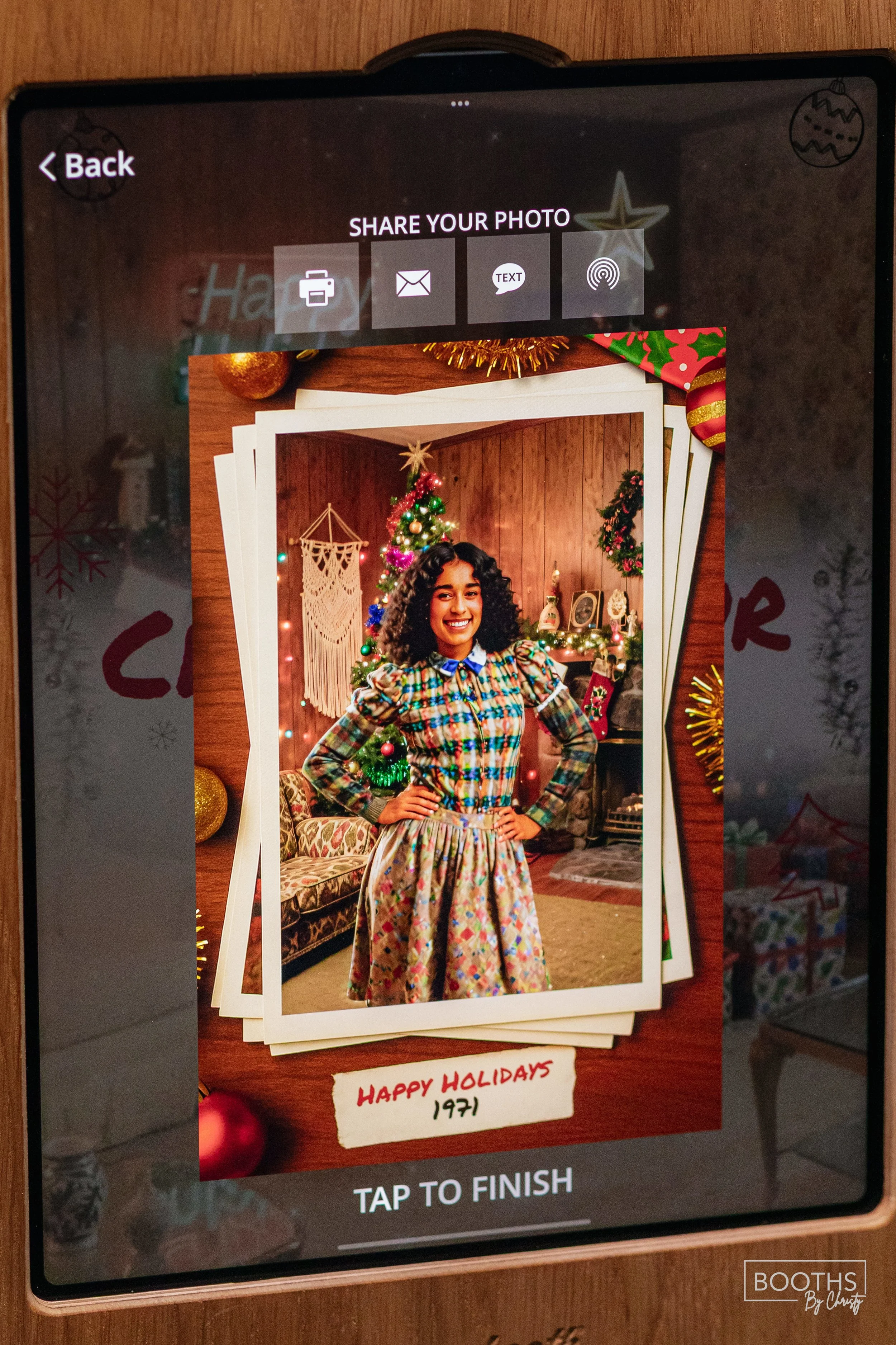 A woman smiling in front of a decorated Christmas tree, wearing a colorful plaid dress, with holiday decor in the background.
