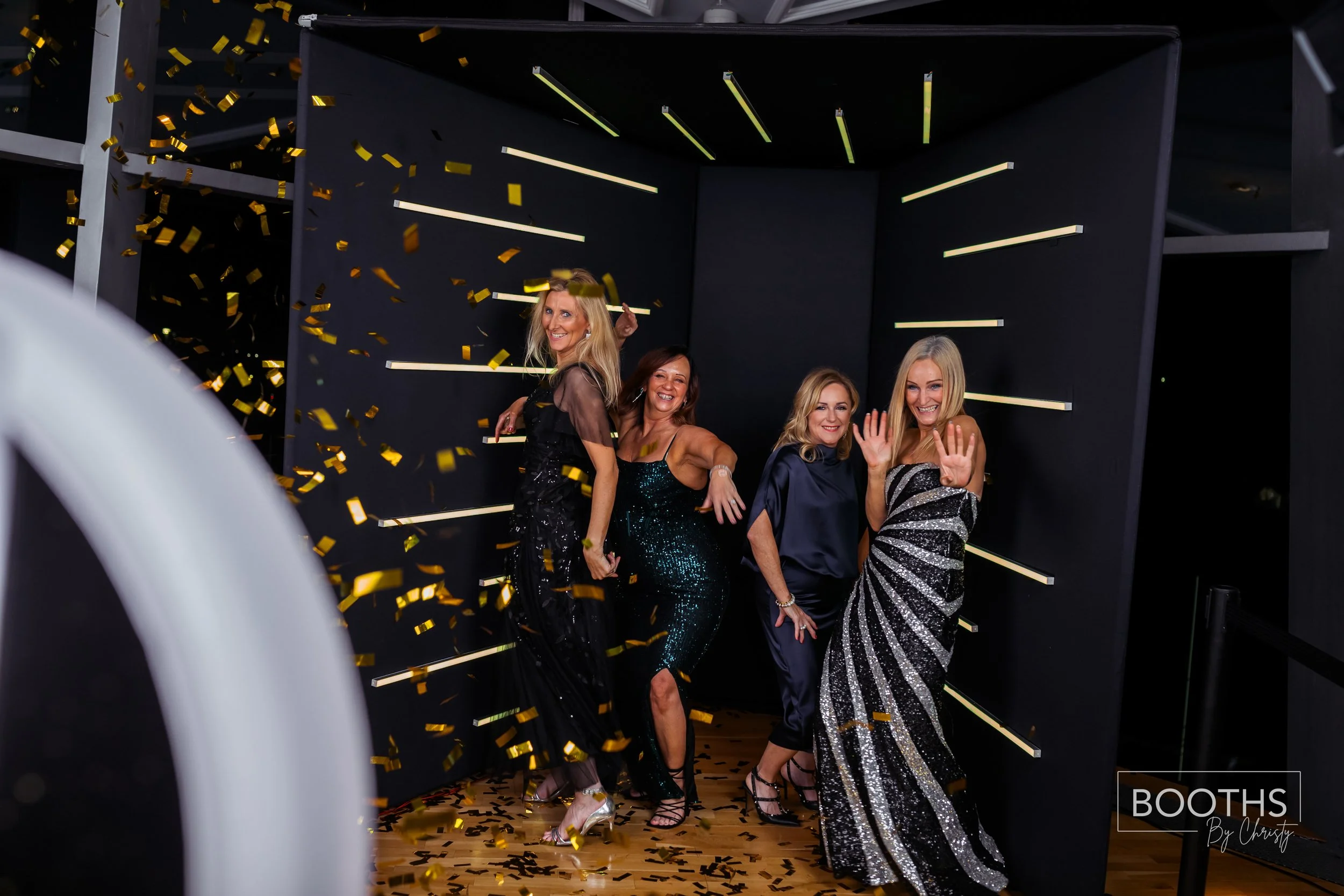 Four women in glamorous dresses celebrate at a photo booth with black walls and gold confetti falling, smiling and posing for the camera.