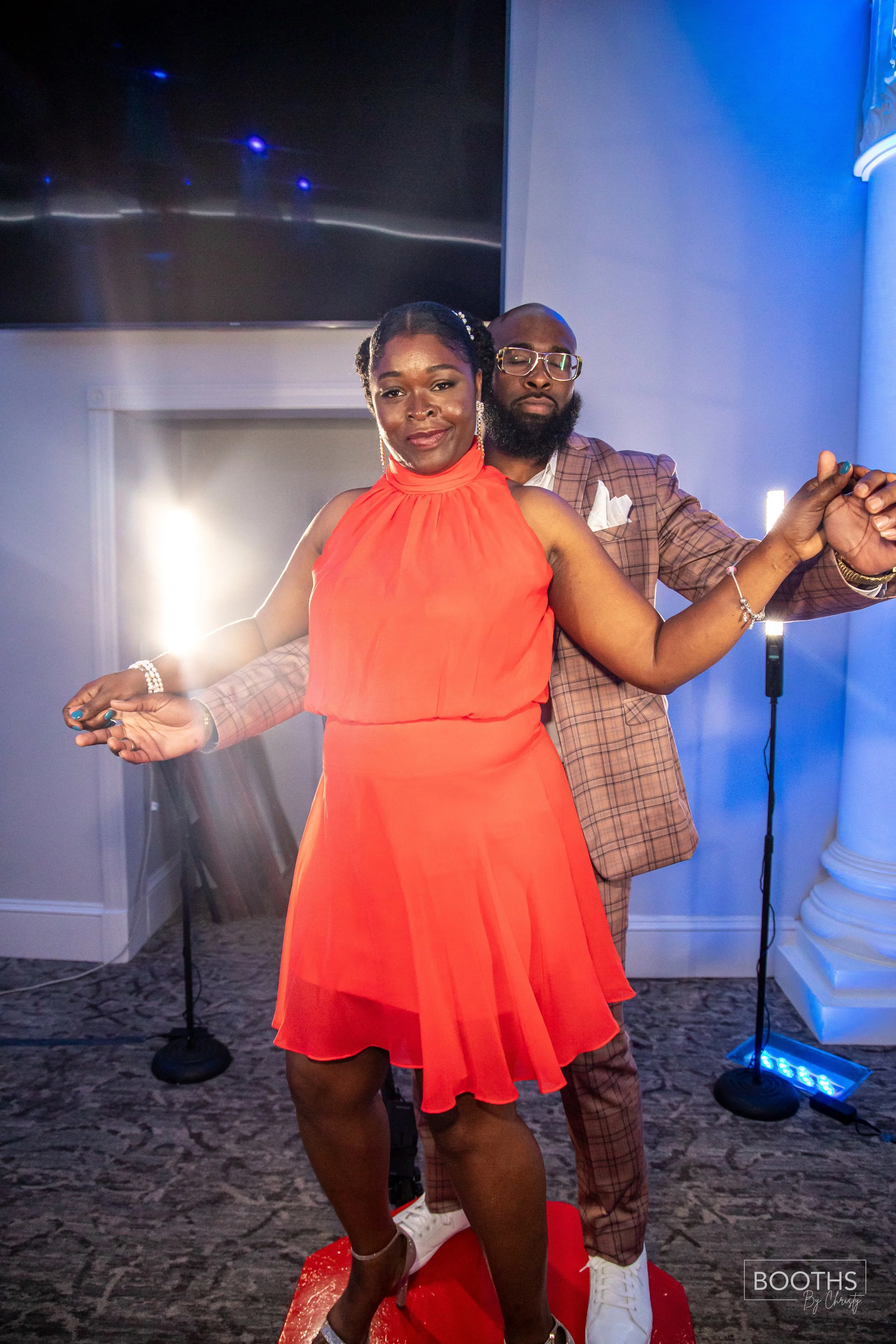 A man and woman dancing together at an indoor event, with the woman in a bright red dress and the man in a checkered suit, both smiling and holding hands.