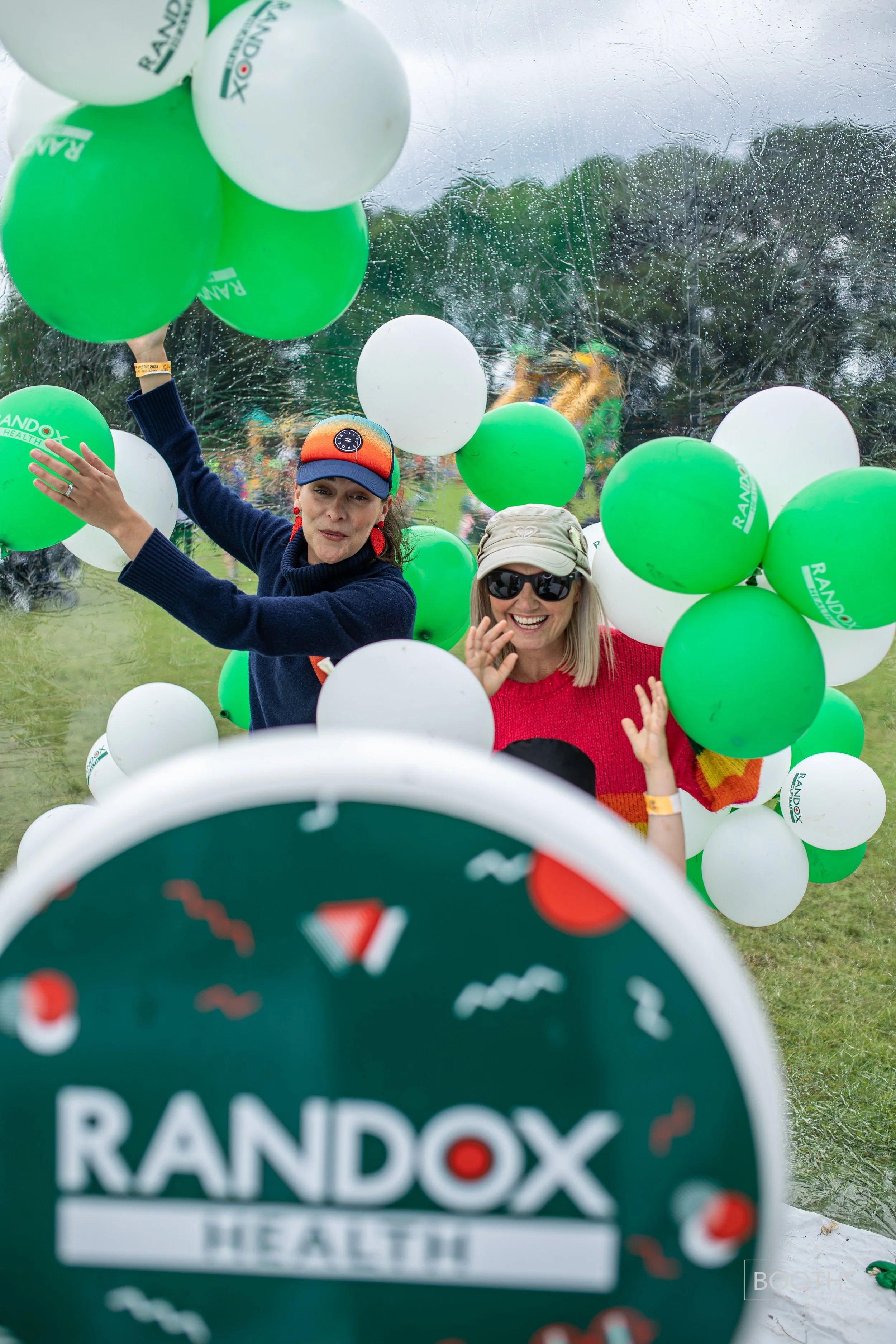 Two women smiling and celebrating at an outdoor event with white and green balloons. The foreground shows a blurred green circular sign with the word 'RANDOX' and the background features trees and a cloudy sky.