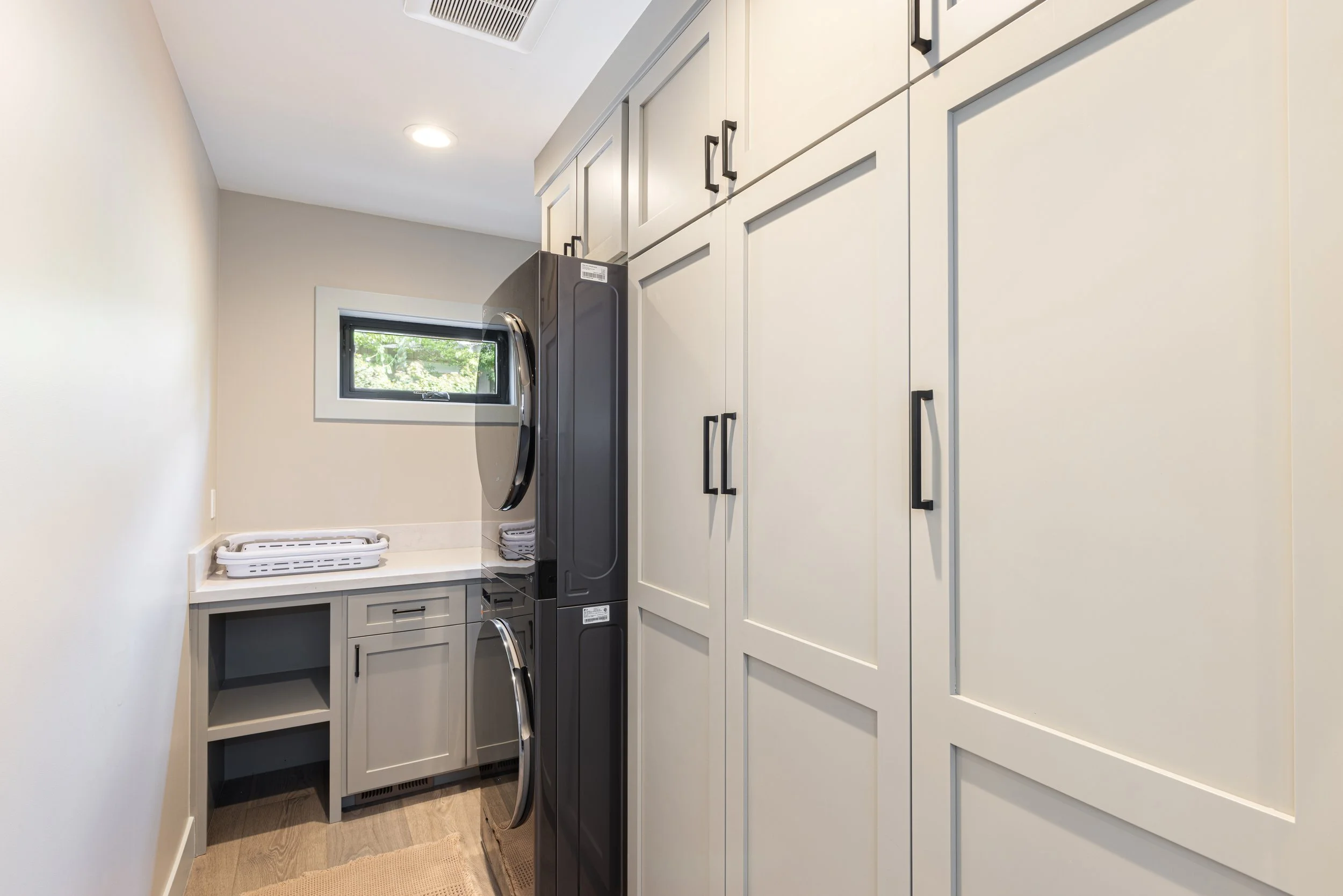 A modern laundry room with a front-loading washer and dryer, white cabinets with black handles, a small window, and a countertop with laundry baskets.