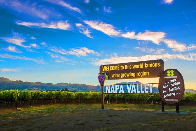 Sign welcoming visitors to Napa Valley, a famous wine-growing region, with vineyards and hills in the background under a partly cloudy sky.