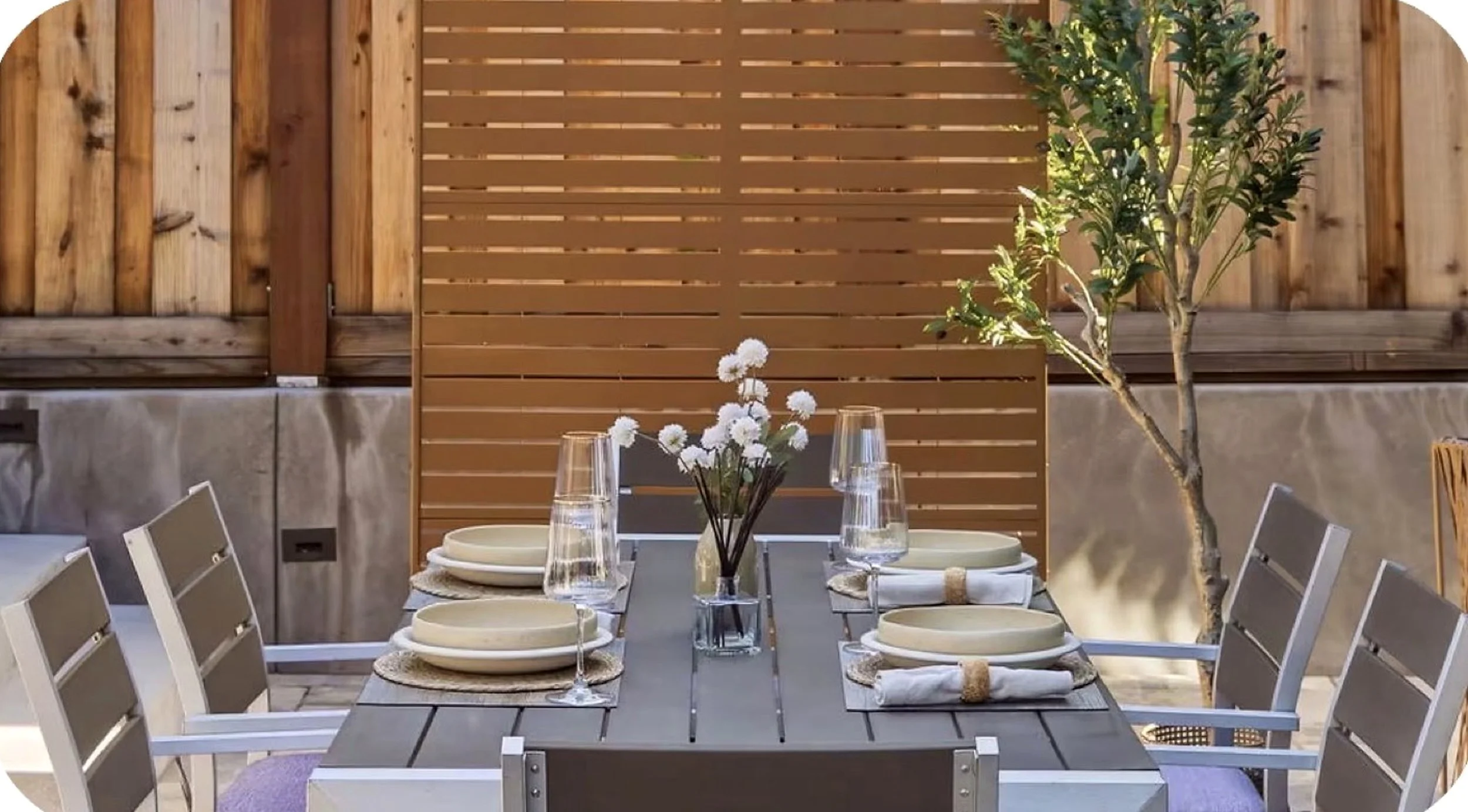 Outdoor dining table set with six beige plates, clear glasses, and a small flower arrangement in a vase. The table is surrounded by white and beige chairs, with a wooden privacy fence and a small tree in the background, creating a cozy patio setting.