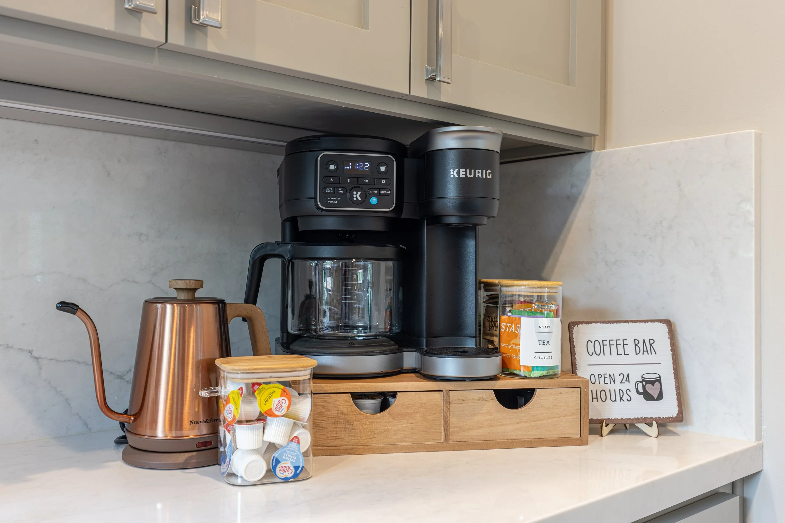 Coffee station with a black Keurig coffee maker, copper electric kettle, small container of creamers, and a sign that reads 'Coffee Bar Open 24 Hours' on a white countertop.