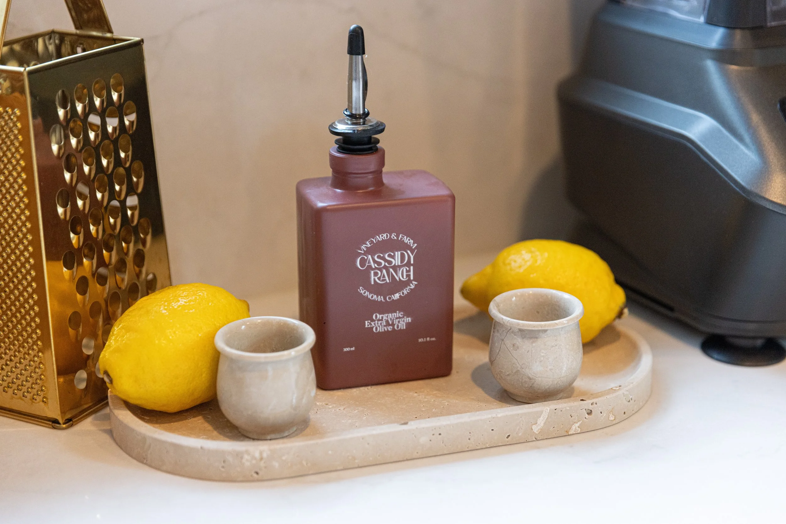 A kitchen countertop tray holds two lemons, two small beige cups, a bottle of Cassidy Rancji olive oil, a lemon, a golden cheese grater, and a black kitchen appliance.