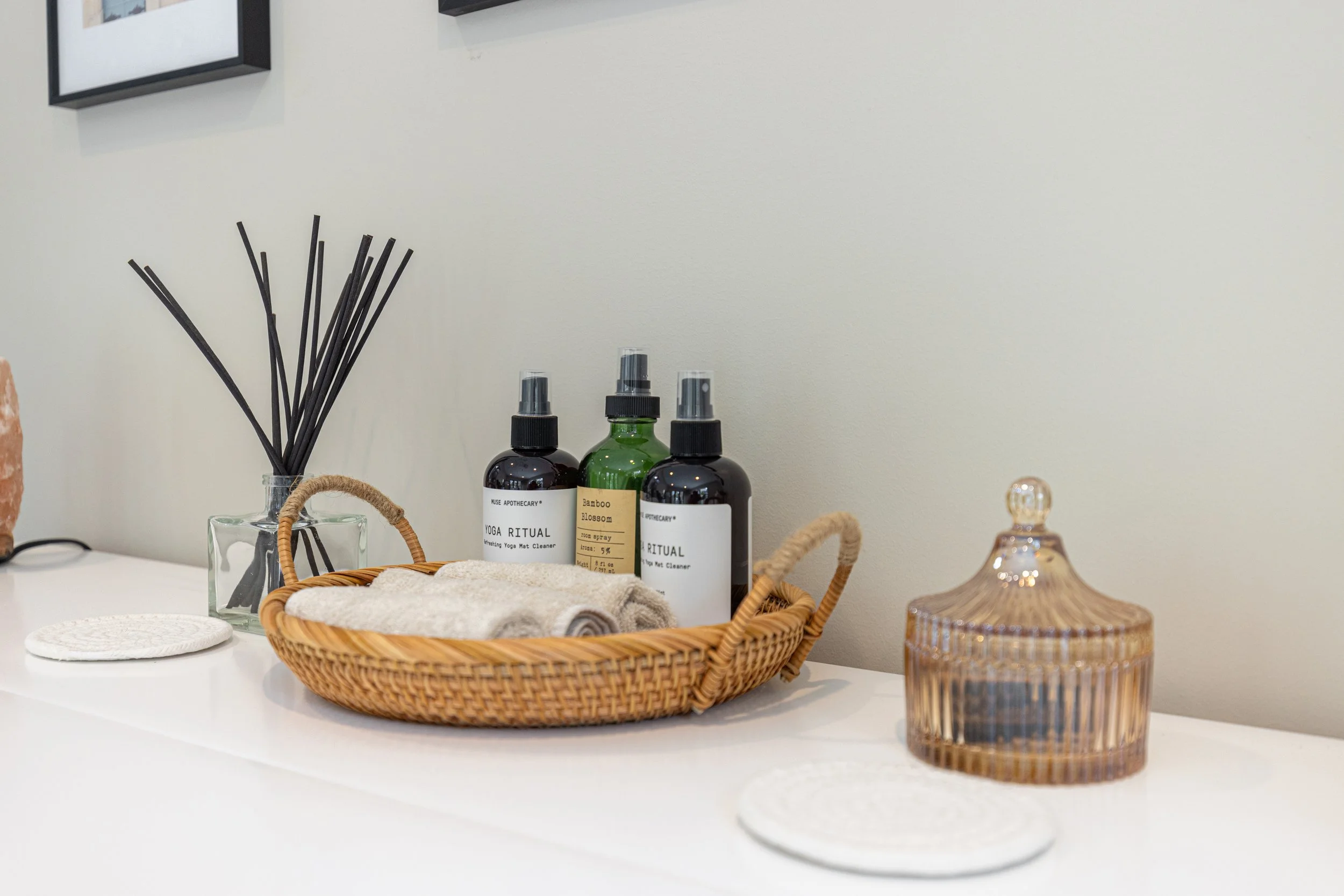 A white surface with a wicker basket holding rolled beige towels and three spray bottles, a reed diffuser in a glass container, a decorative container with a lid, and two round white coasters.