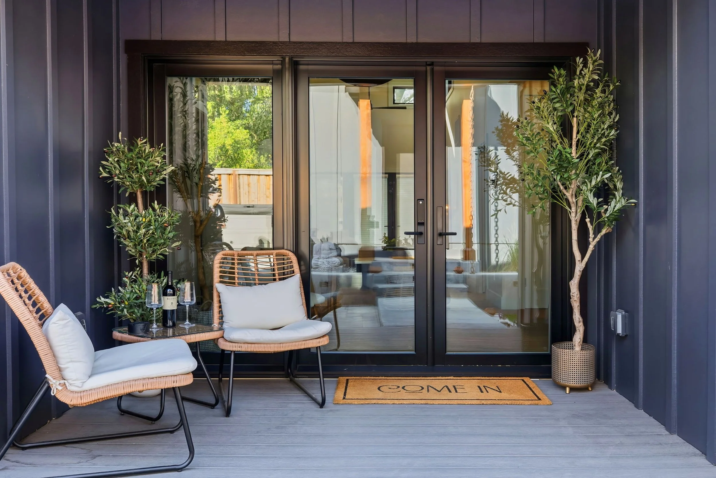 A cozy outdoor patio with wicker chairs, white cushions, a small table set with wine glasses and a bottle, potted plants, a welcome mat, and glass sliding doors leading into a house.