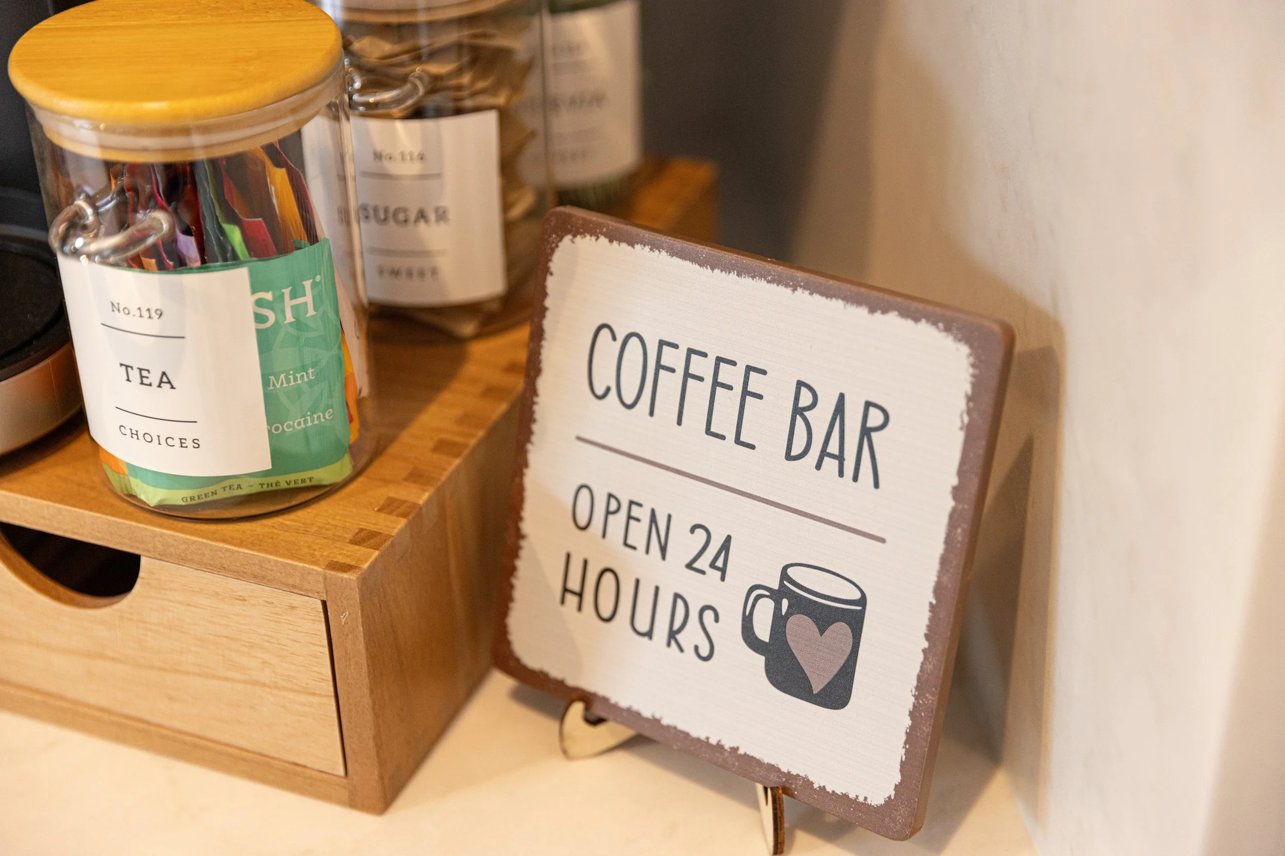 A sign on a stand reading 'Coffee Bar, Open 24 Hours' with a coffee mug illustration, placed on a countertop next to jars of tea and sugar.