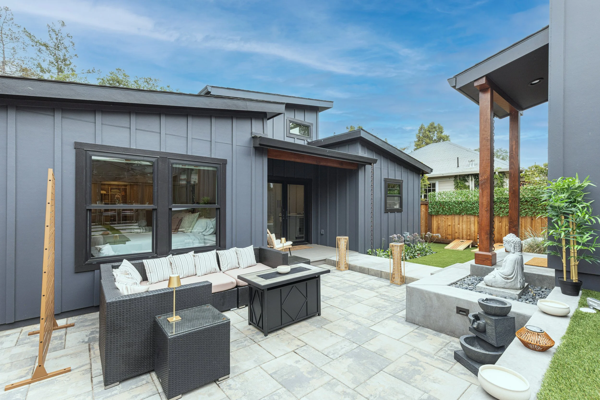 Modern backyard patio with outdoor sofa, coffee table, Buddha statue, decorative stones, plants, and a wooden fence, under a blue sky.