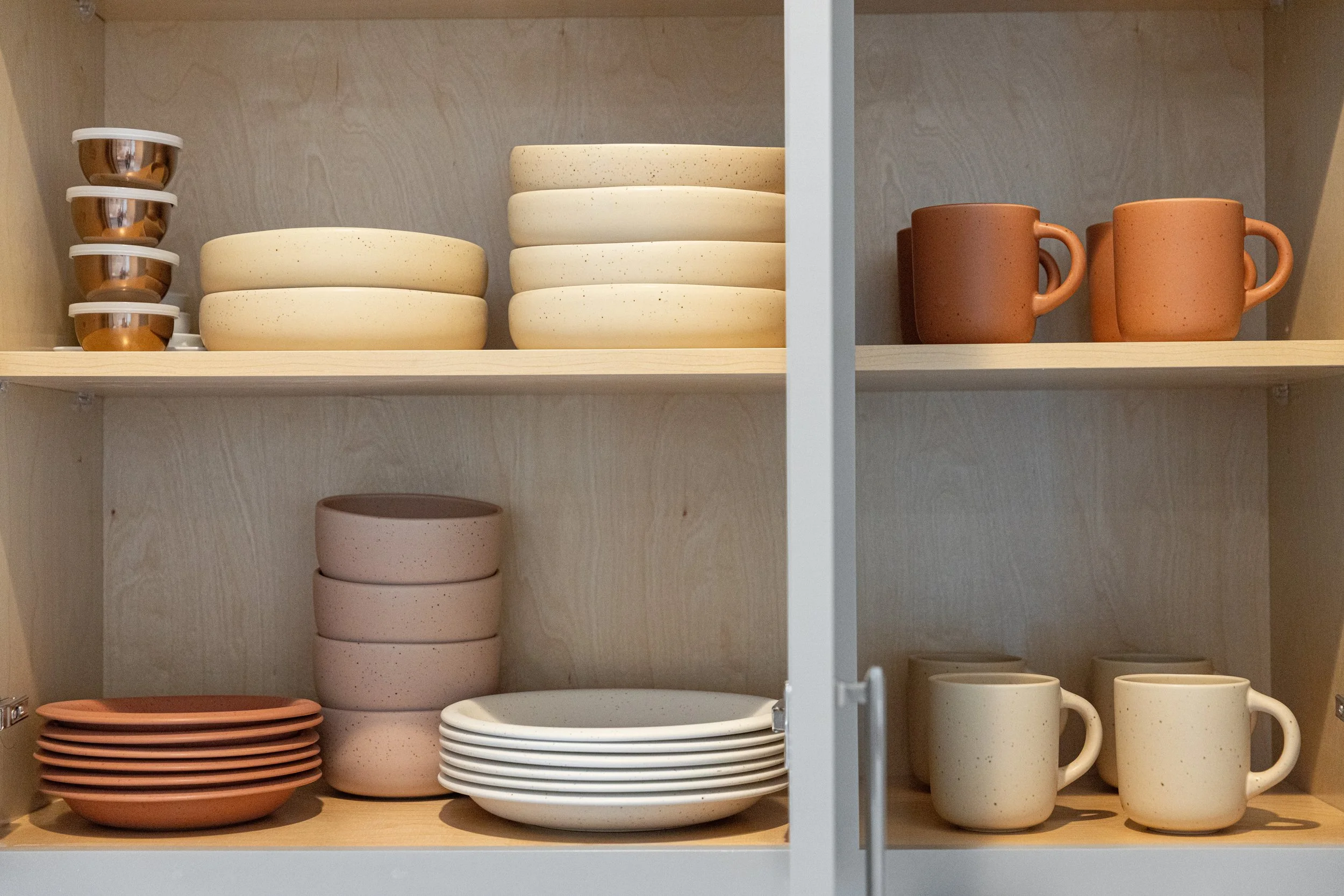 Open kitchen cabinet with stacked cream-colored bowls and small terracotta cups on the top shelf, pink and white plates with matching bowls on the bottom shelf, and cream-colored coffee mugs on the bottom right shelf.