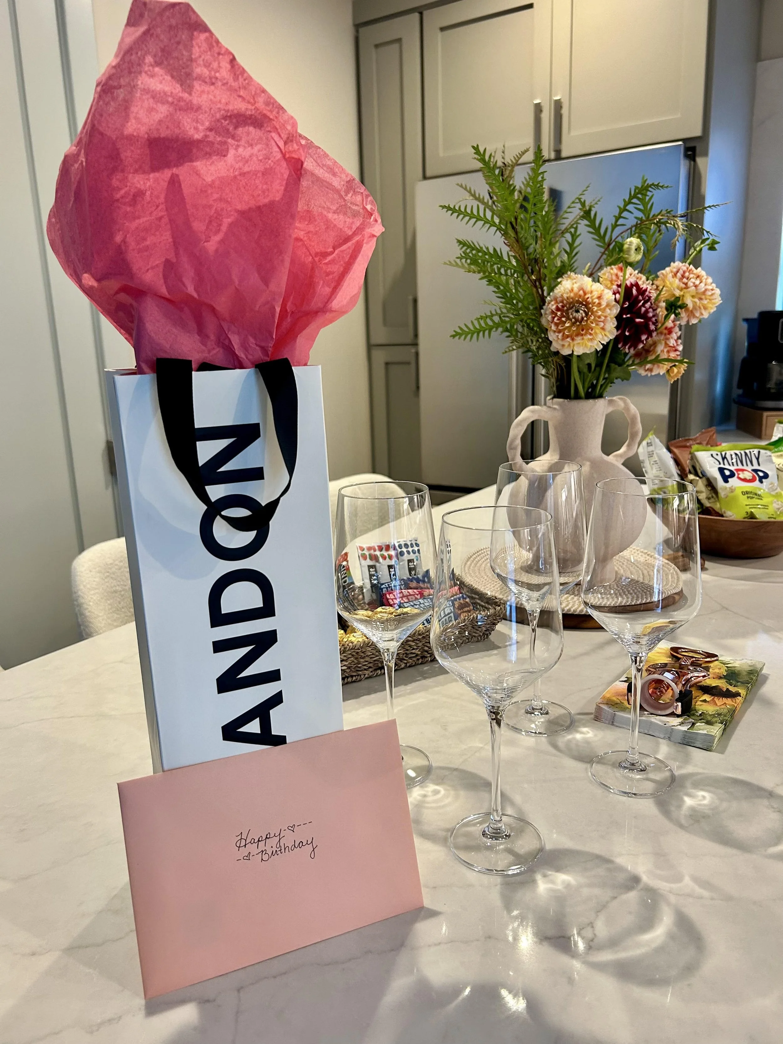 Table setting with a white Amani shopping bag containing pink tissue paper, four empty wine glasses, a pink birthday card, a vase with pink and yellow flowers and green foliage, snacks, and a decorative woven tray.