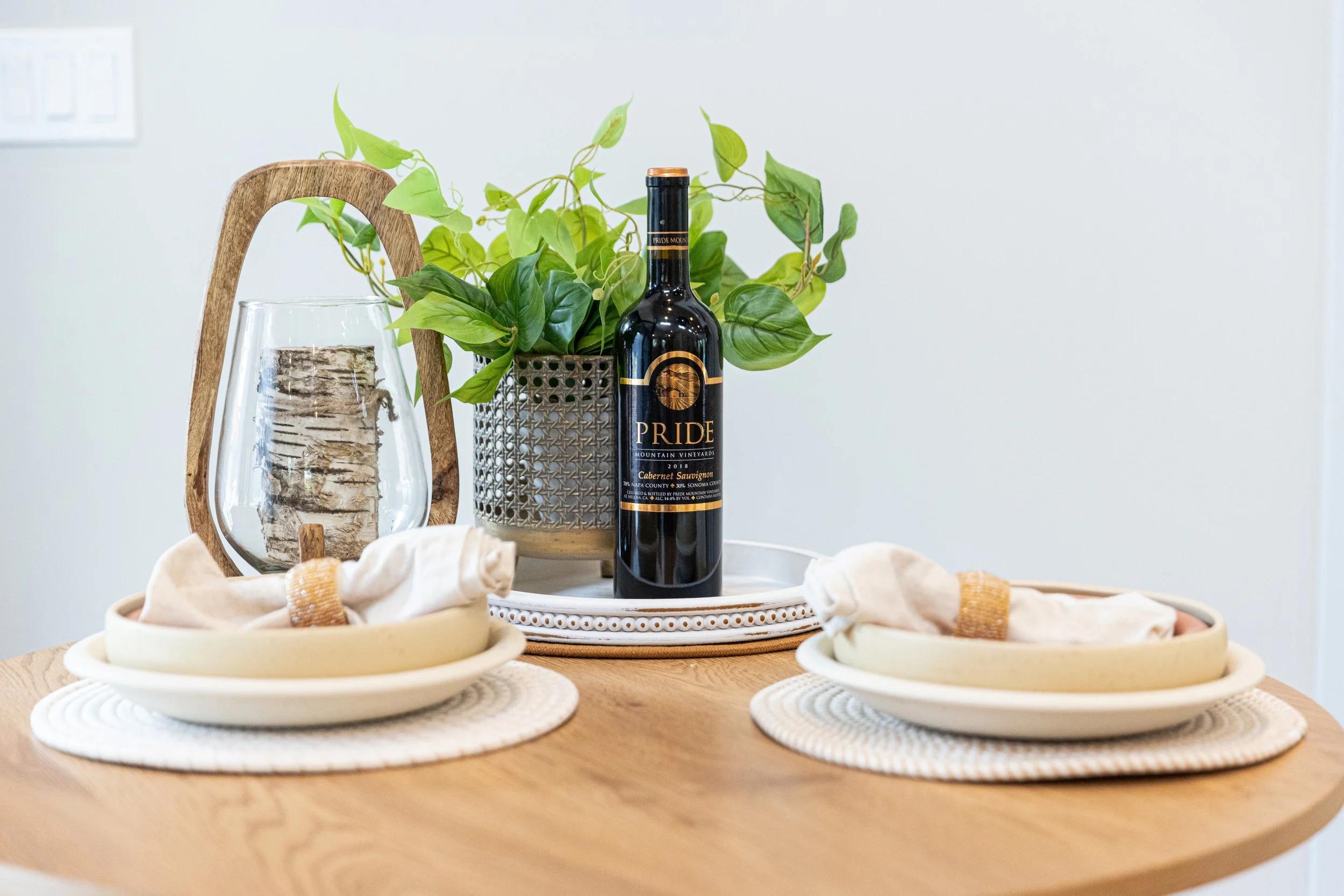 A dining table with two place settings, folded napkins, a bottle of wine labeled 'Pride,' a decorative glass container with a log inside, and a potted plant in a textured planter.