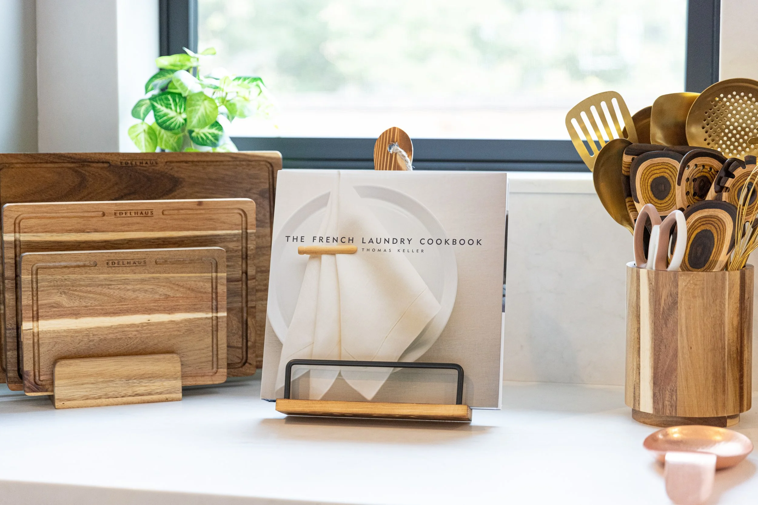 Kitchen countertop with wooden cutting boards, a cookbook titled "The French Laundry Cookbook" by Thomas Keller, and a container holding wooden utensils with various handles near a window with a green plant in the background.