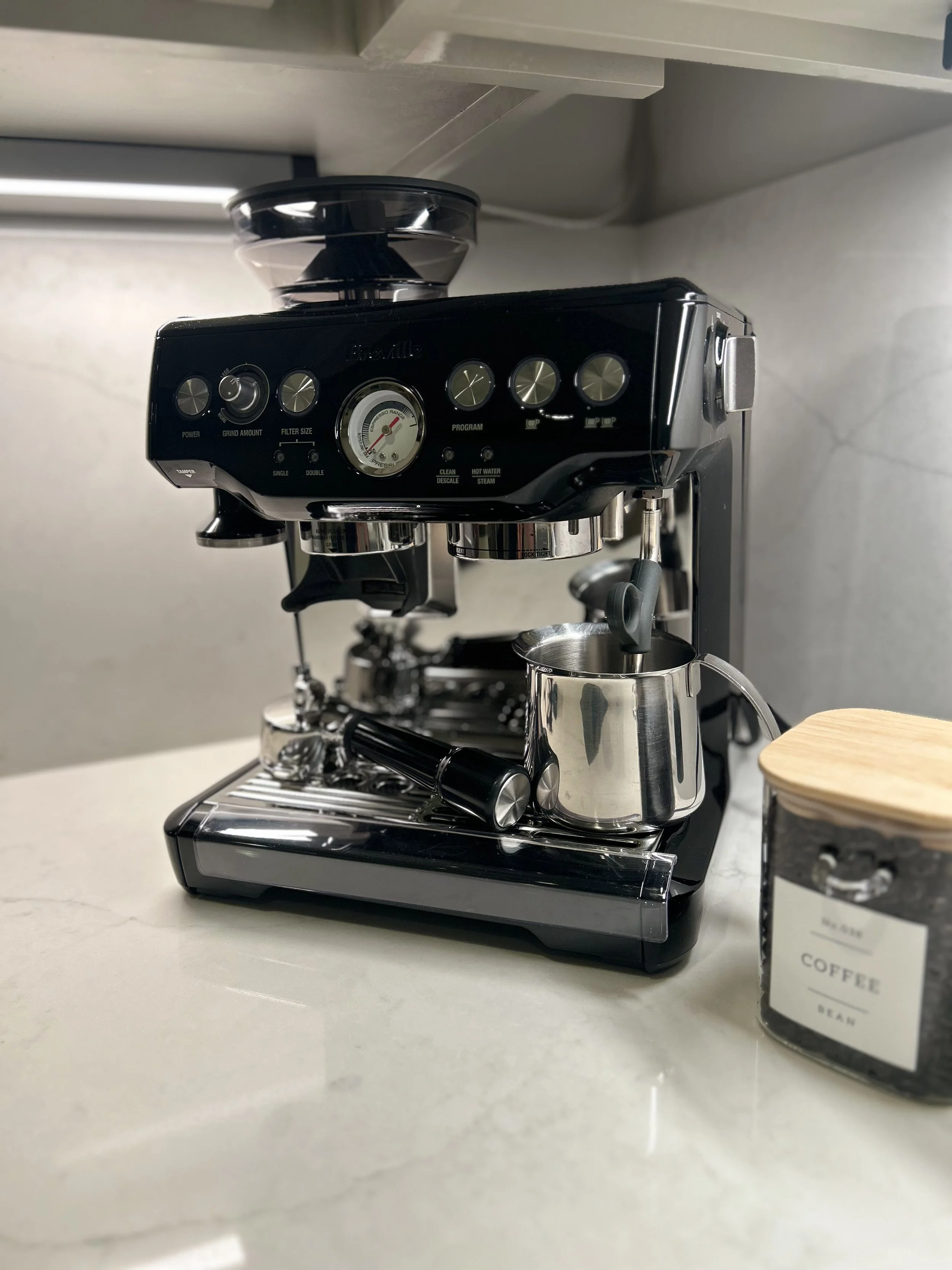 Black espresso machine on kitchen counter, with a small stainless steel pitcher, a coffee tamper, and a jar labeled 'Coffee Bean' nearby.