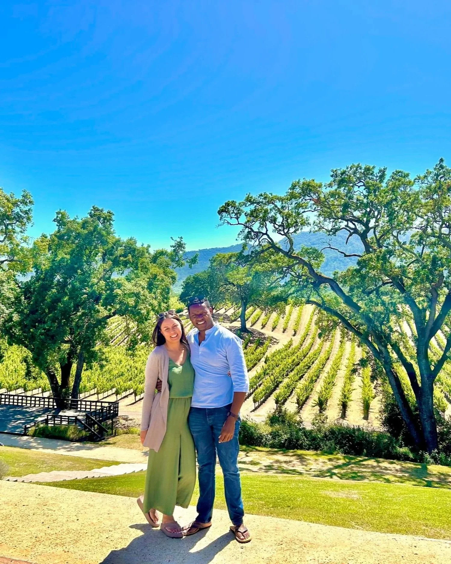 A smiling couple standing together outdoors in a vineyard with green trees and rolling hills in the background on a sunny day.