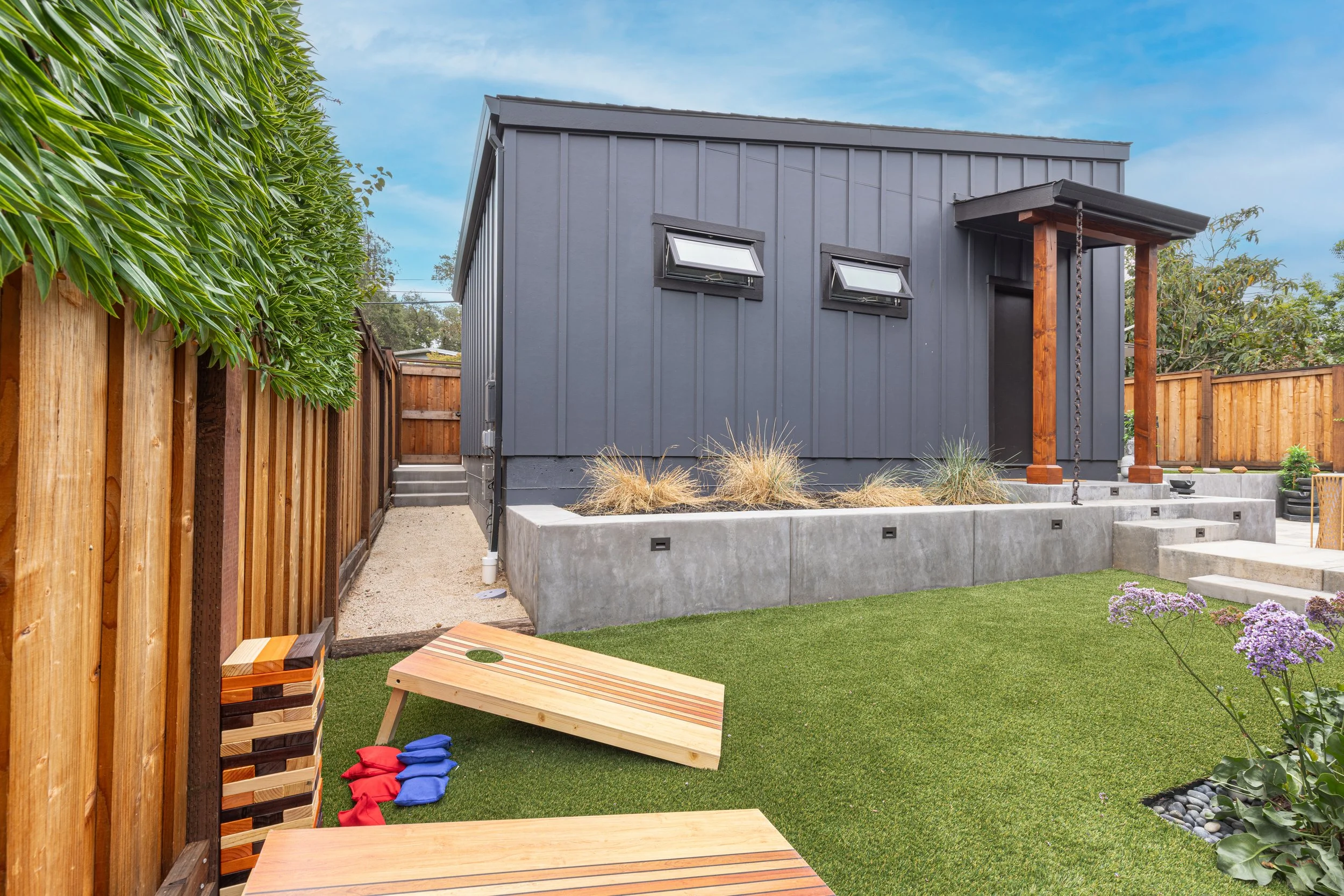 A modern backyard with a dark gray house, small windows, a wooden porch with a chain hanging from the roof, a concrete staircase, vibrant green artificial lawn, and a wooden cornhole game with foam gloves nearby.