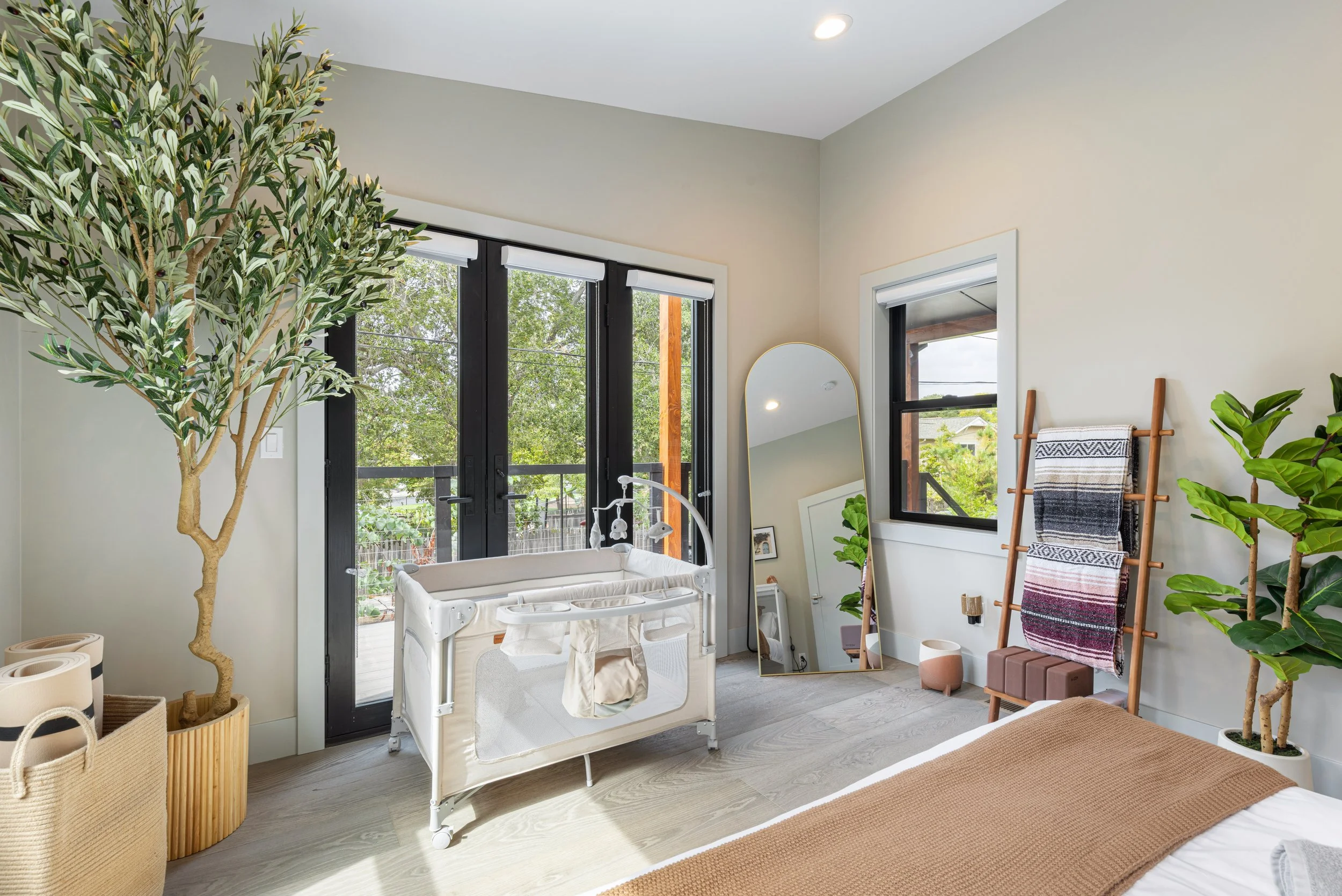 A bright bedroom with large windows, a mirror, a crib, plants, and a blanket rack.