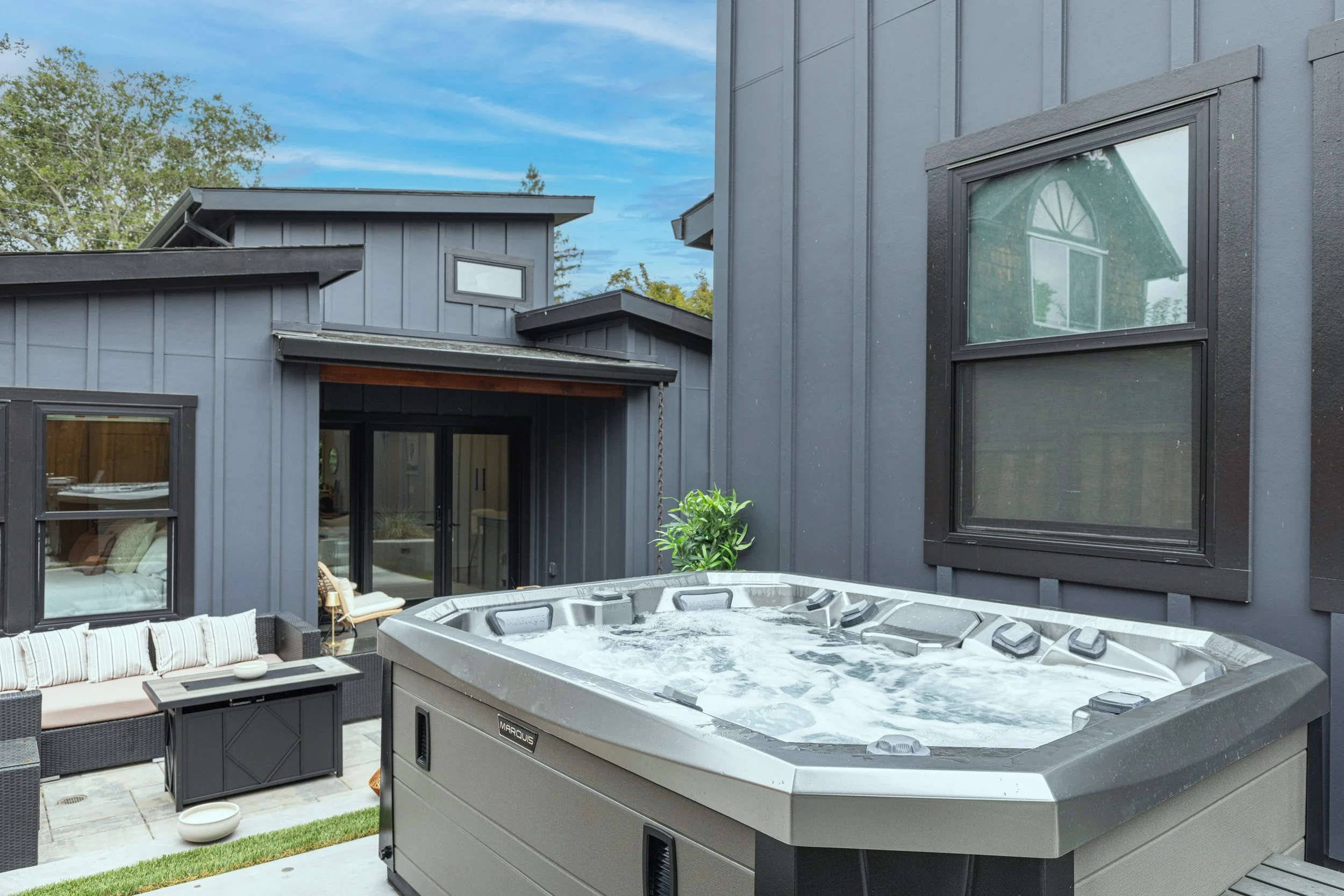 Outdoor patio area featuring a hot tub, black wicker outdoor furniture with white cushions, a small storage bench, and black-framed windows and sliding doors on modern gray house exterior, with blue sky overhead.