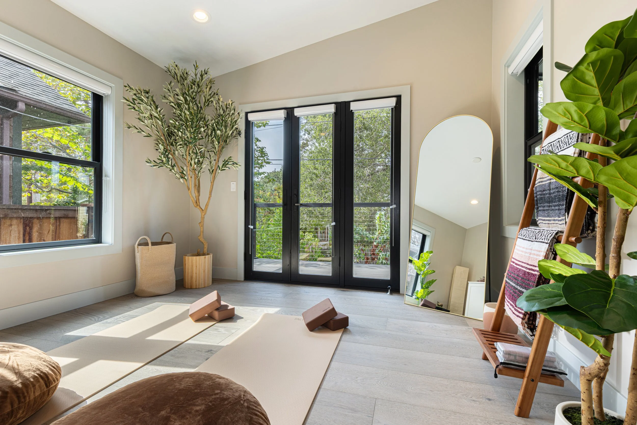 Bright airy room with large windows, a potted tree, yoga mats with blocks, a mirror resting on the floor, and a wooden ladder with towels, decorated in neutral tones and natural materials.
