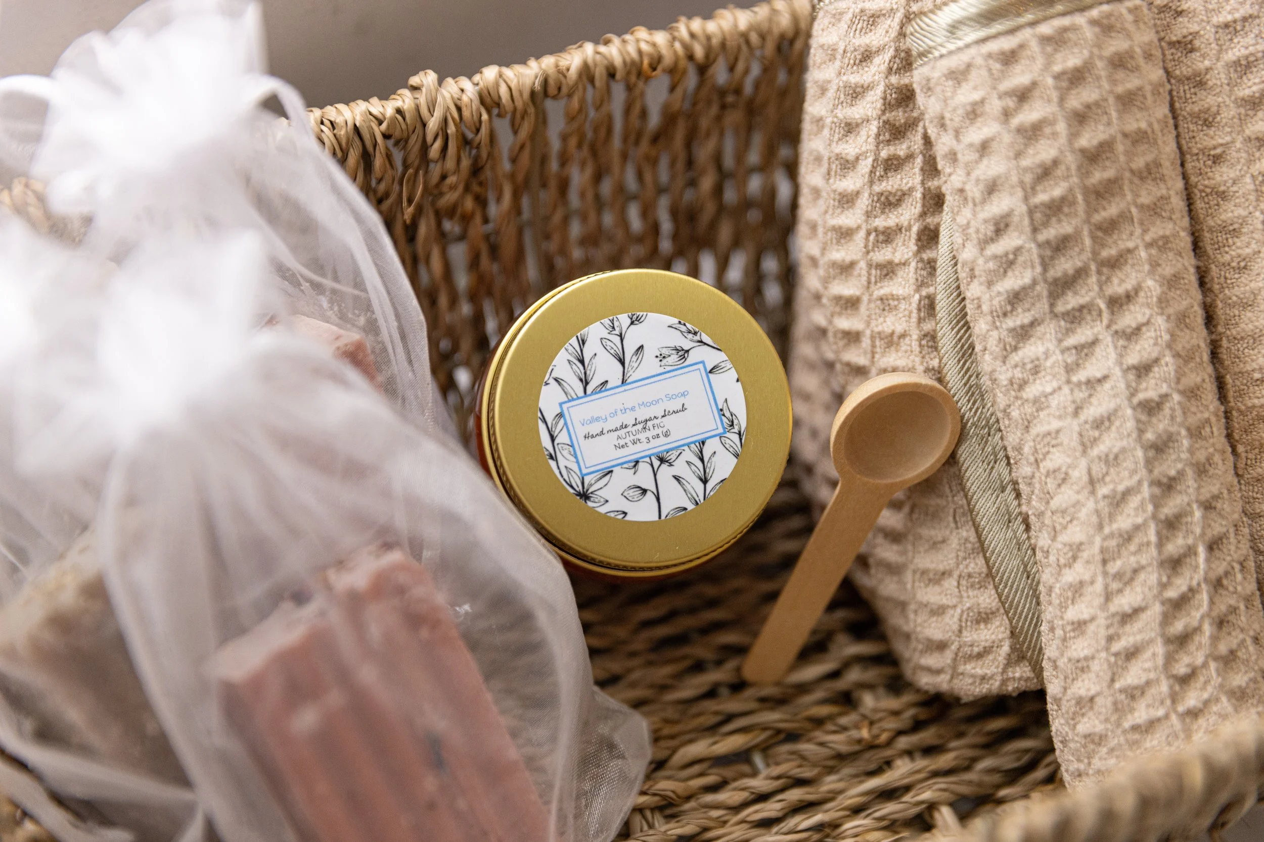 A wicker basket containing a jar of handmade soap labeled 'Valley of the Moon Soap,' a wooden scoop, and stacked beige towels.
